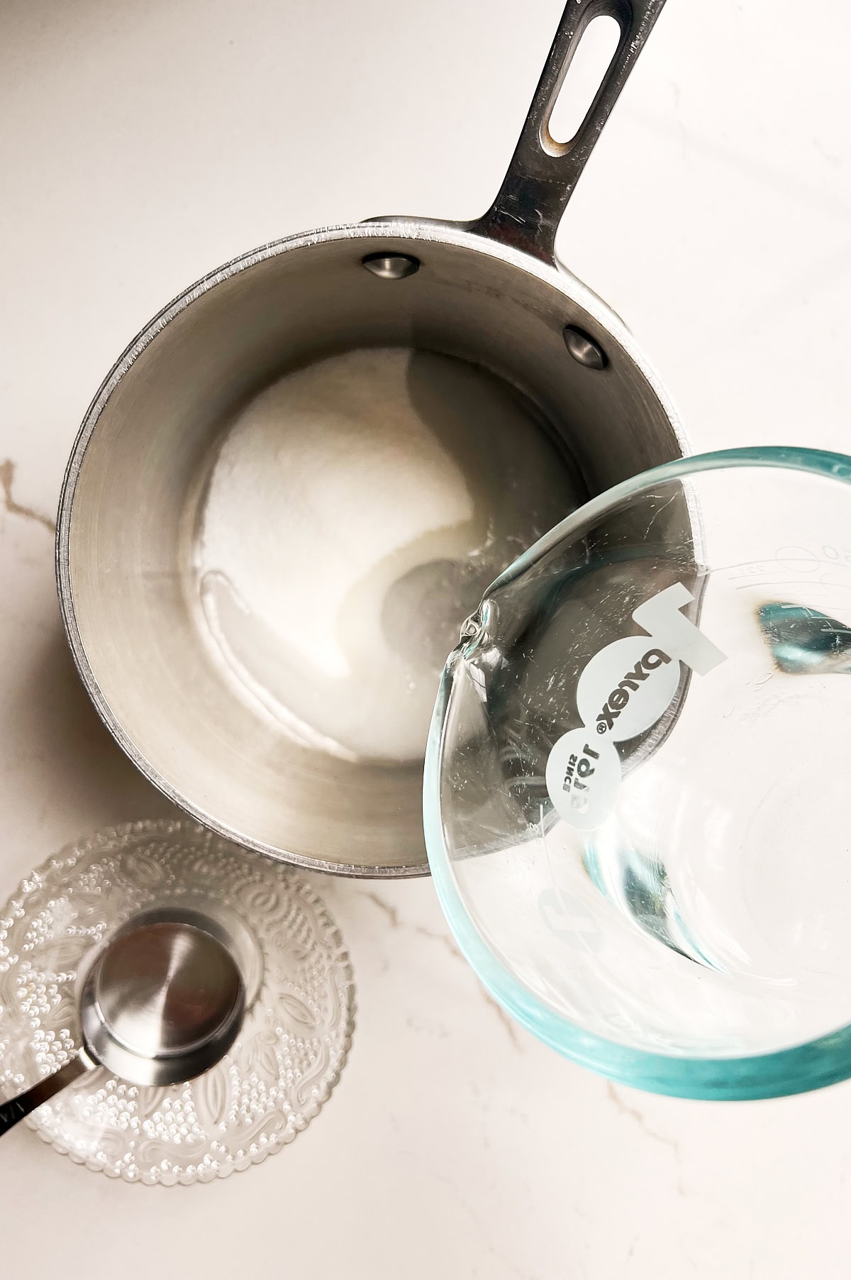 Pyrex of water being poured into a saucepan of sugar on a white counter.