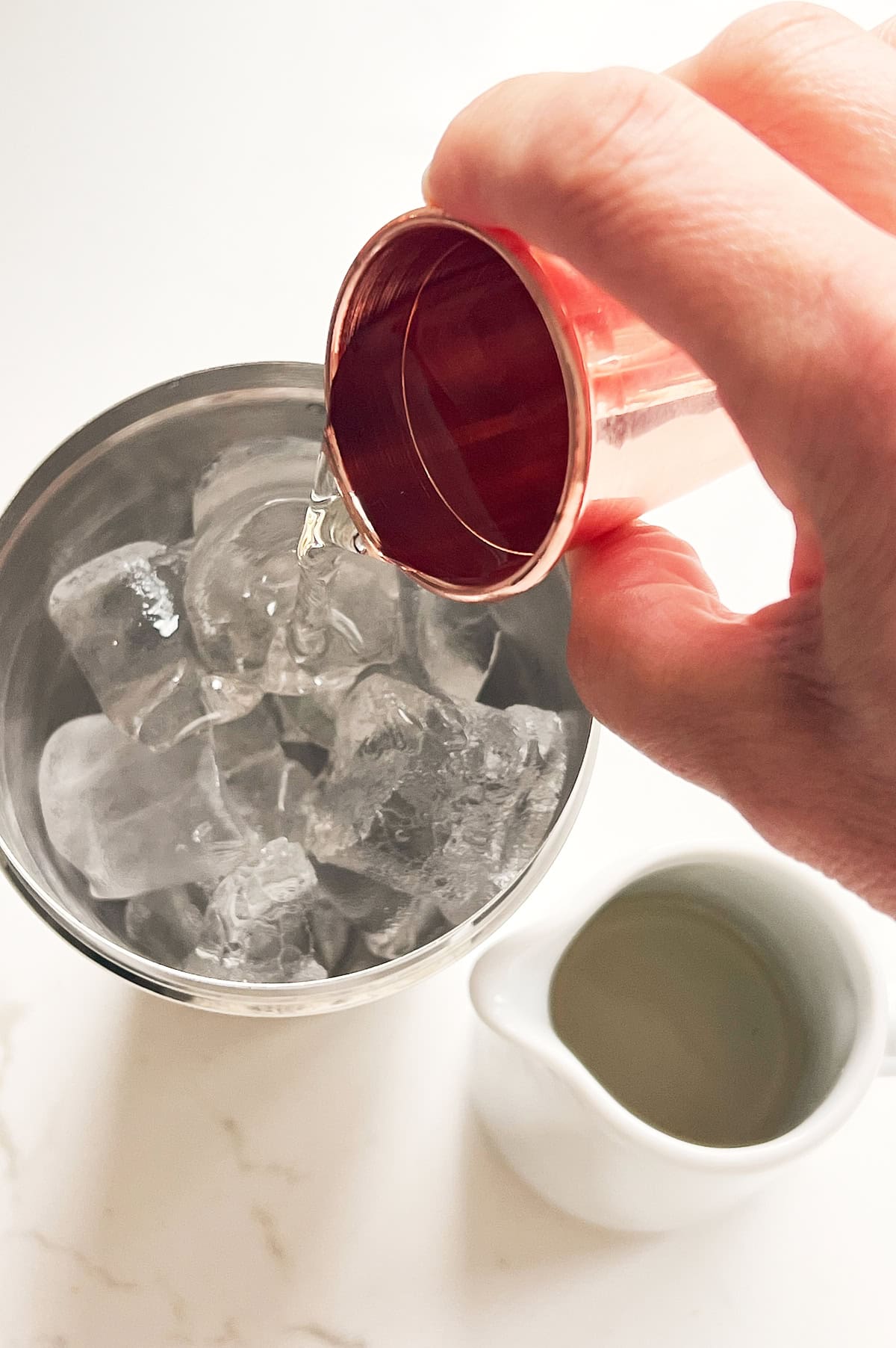 Hand pouring simple syrup over ice in a cocktail shaker on a white counter.