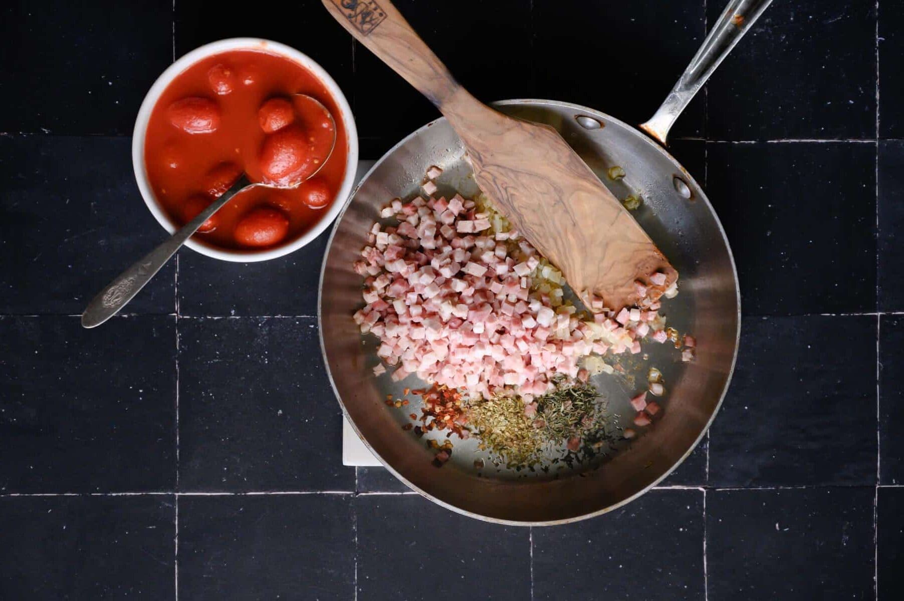 skillet with prosciutto, seasonings and a white bowl with canned tomatoes.