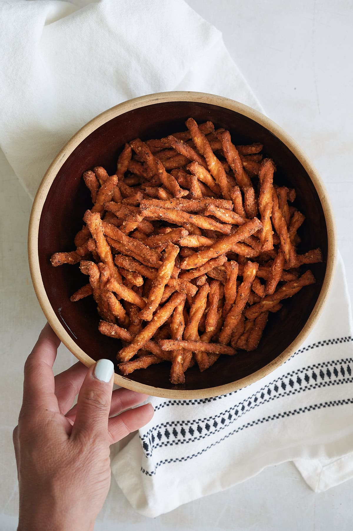 hand reaching for a dark bowl of southwest seasoned pretzels on white napkin with black embroidery.