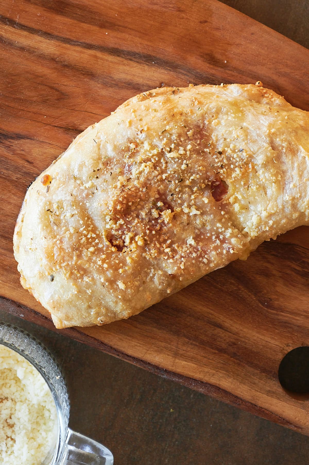 Baked calzone on a wooden board with parmesan and marinara dip nearby.