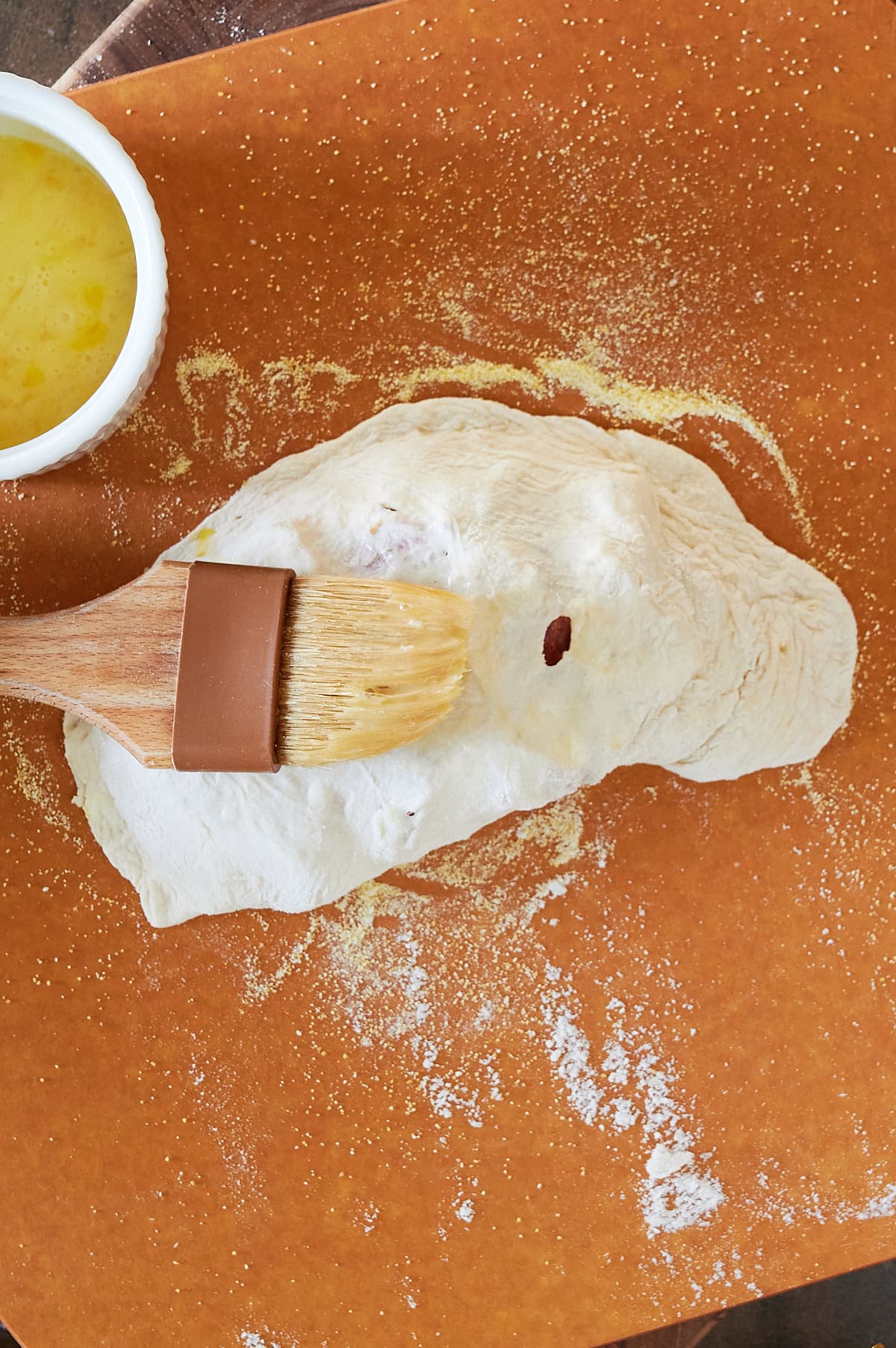 cornmeal sprinkled pizza peel unbaked calzone being brushed with egg.