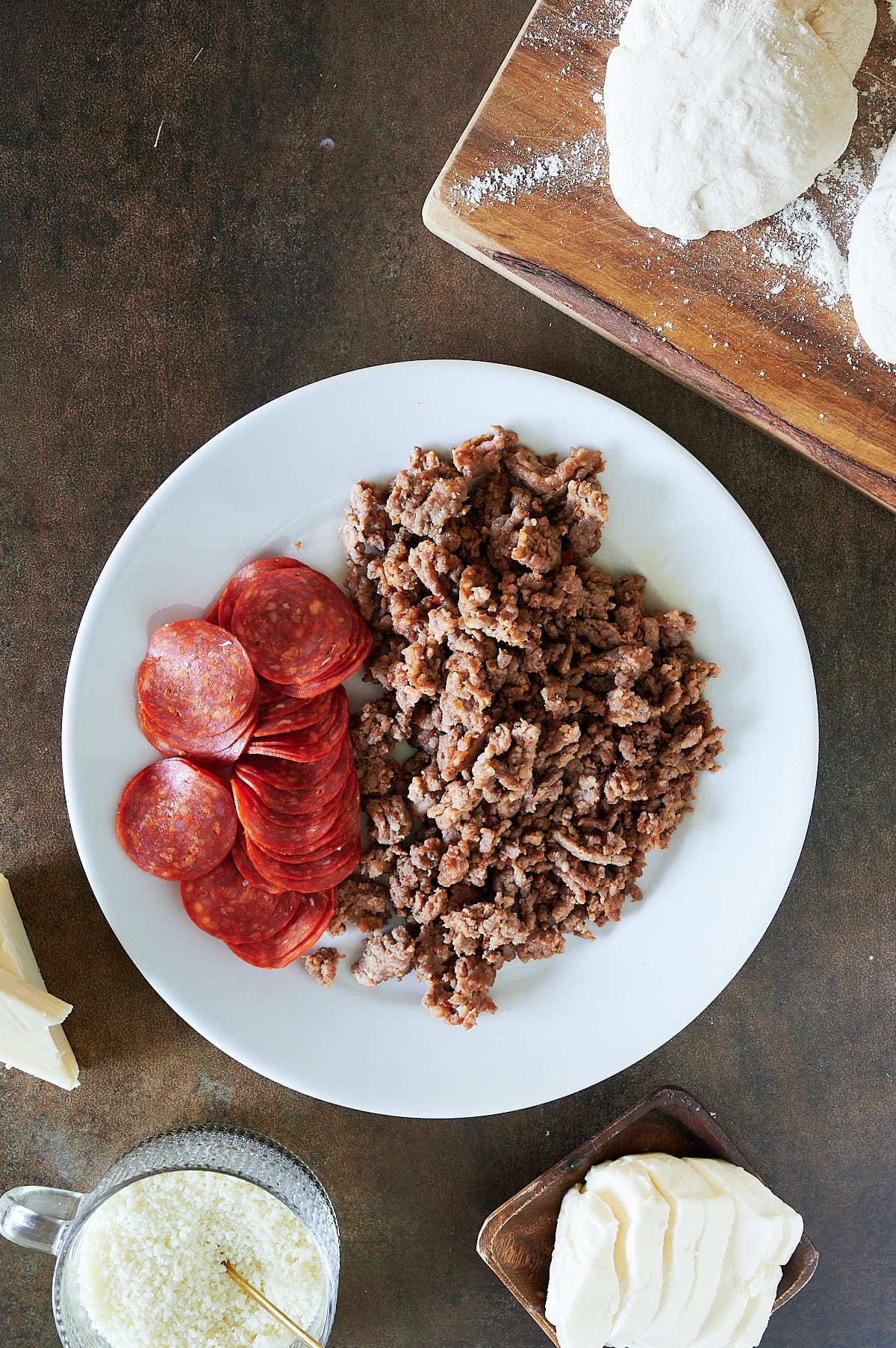 Italian sausage and pepperoni on a white plate with calzone dough and bowls of parmesan and fresh mozzarella.
