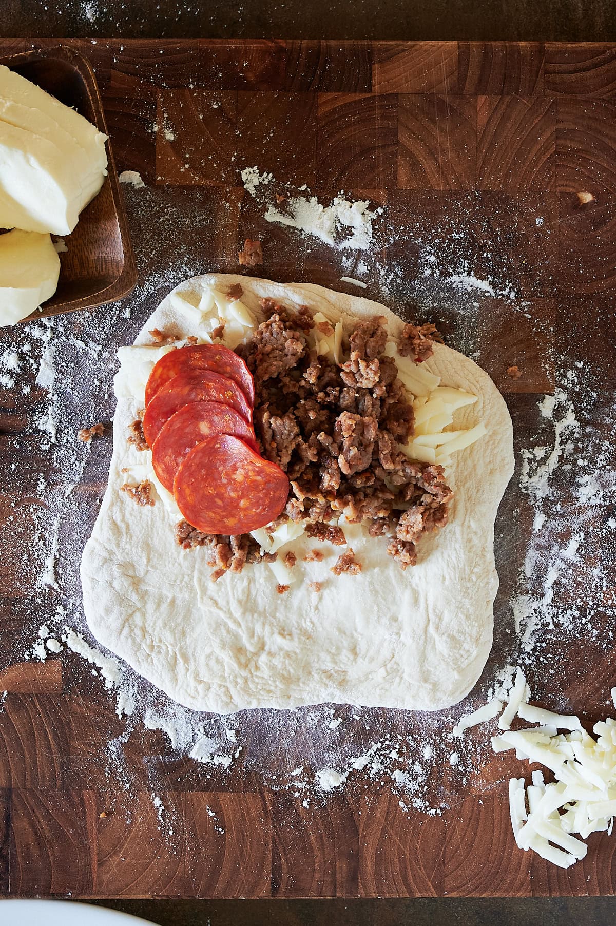 calzone being topped with Italian sausage and pepperoni on a floured wooden board.