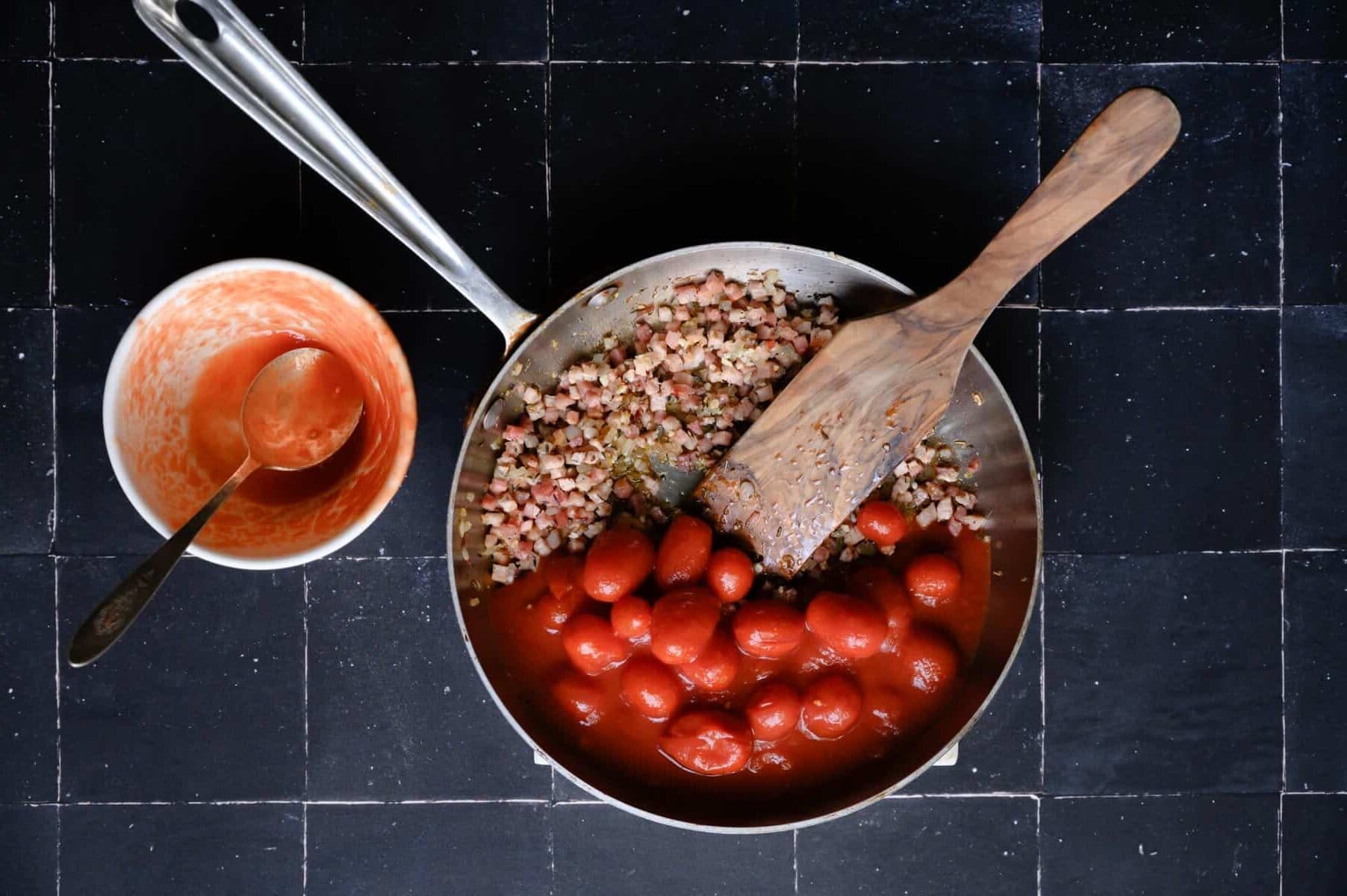 skillet with tomatoes and prosciutto and a wooden paddle.