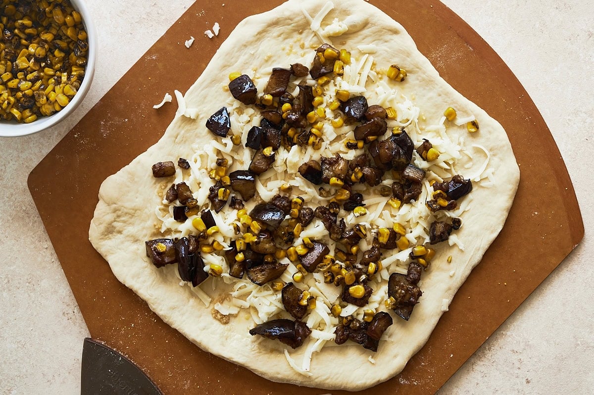 brown pizza peel with an unbaked pizza dough topped with cheese, corn, and eggplant and a bowl of yellow corn nearby.