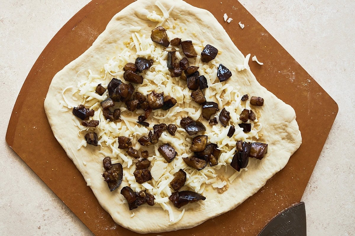 unbaked corn cheese pizza with eggplant on a brown pizza peel and cream background.