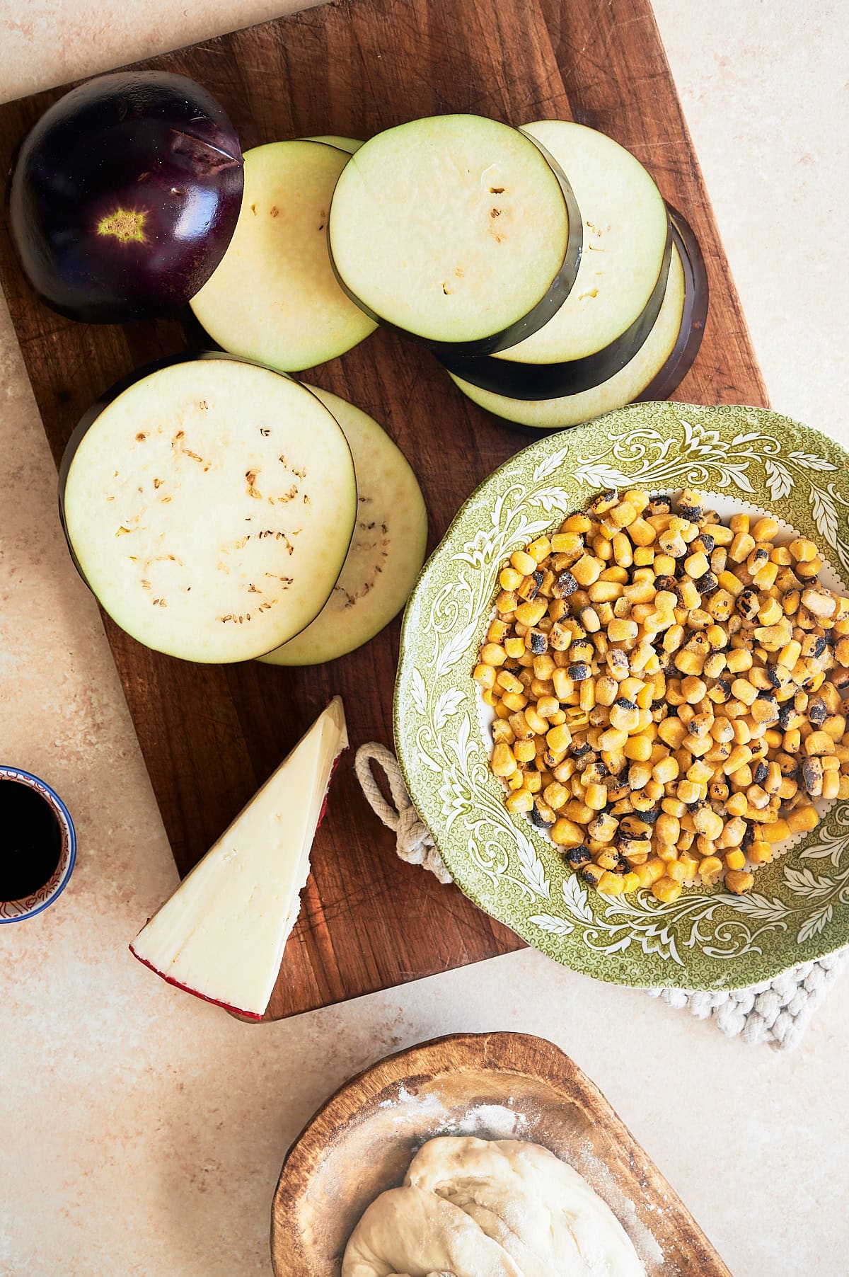Slices of eggplant and a wedge of fontina cheese on a brown cutting board with a bowls of frozen corn, pizza dough and balsamic vinegar nearby.