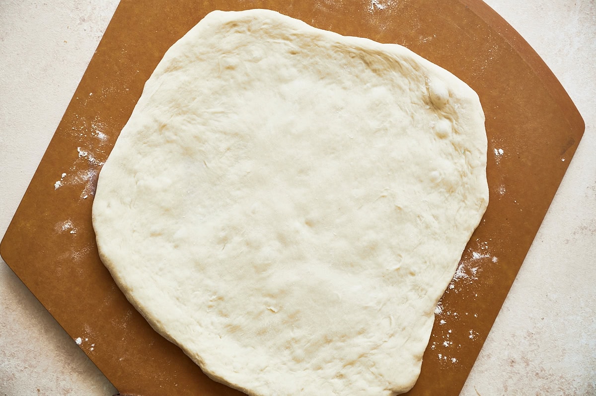 unbaked pizza dough rolled out on brown pizza peel on a cream background.