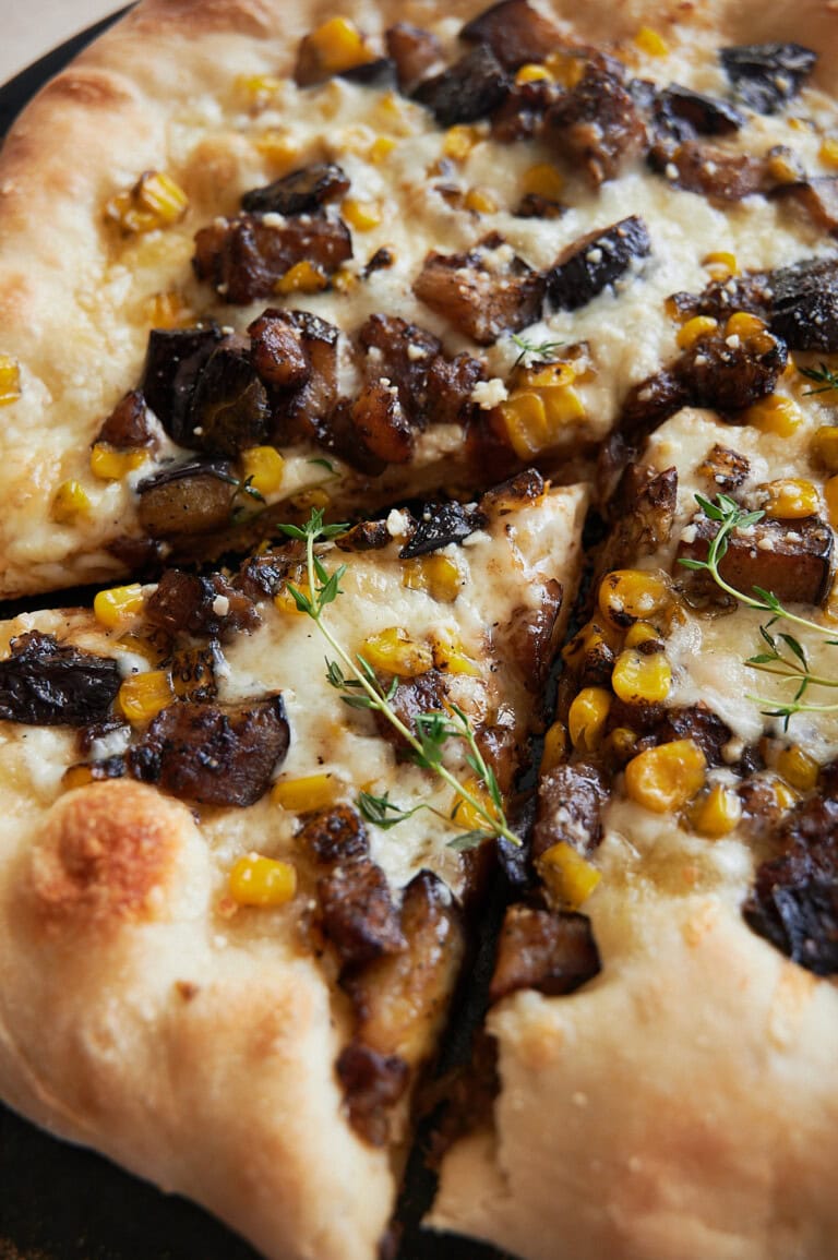 A cut piece of corn cheese pizza with eggplant and fresh thyme on a black pizza stone.