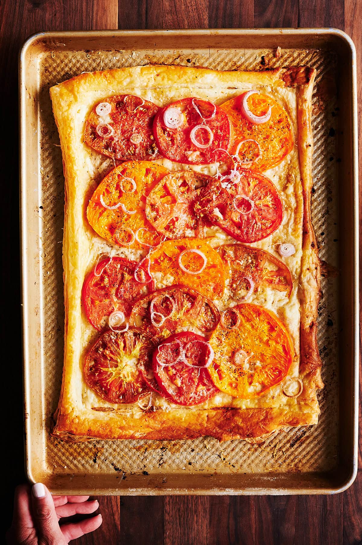 baked heirloom tomato tart with shallots on a sheet pan with a hand placing it on a wooden board.