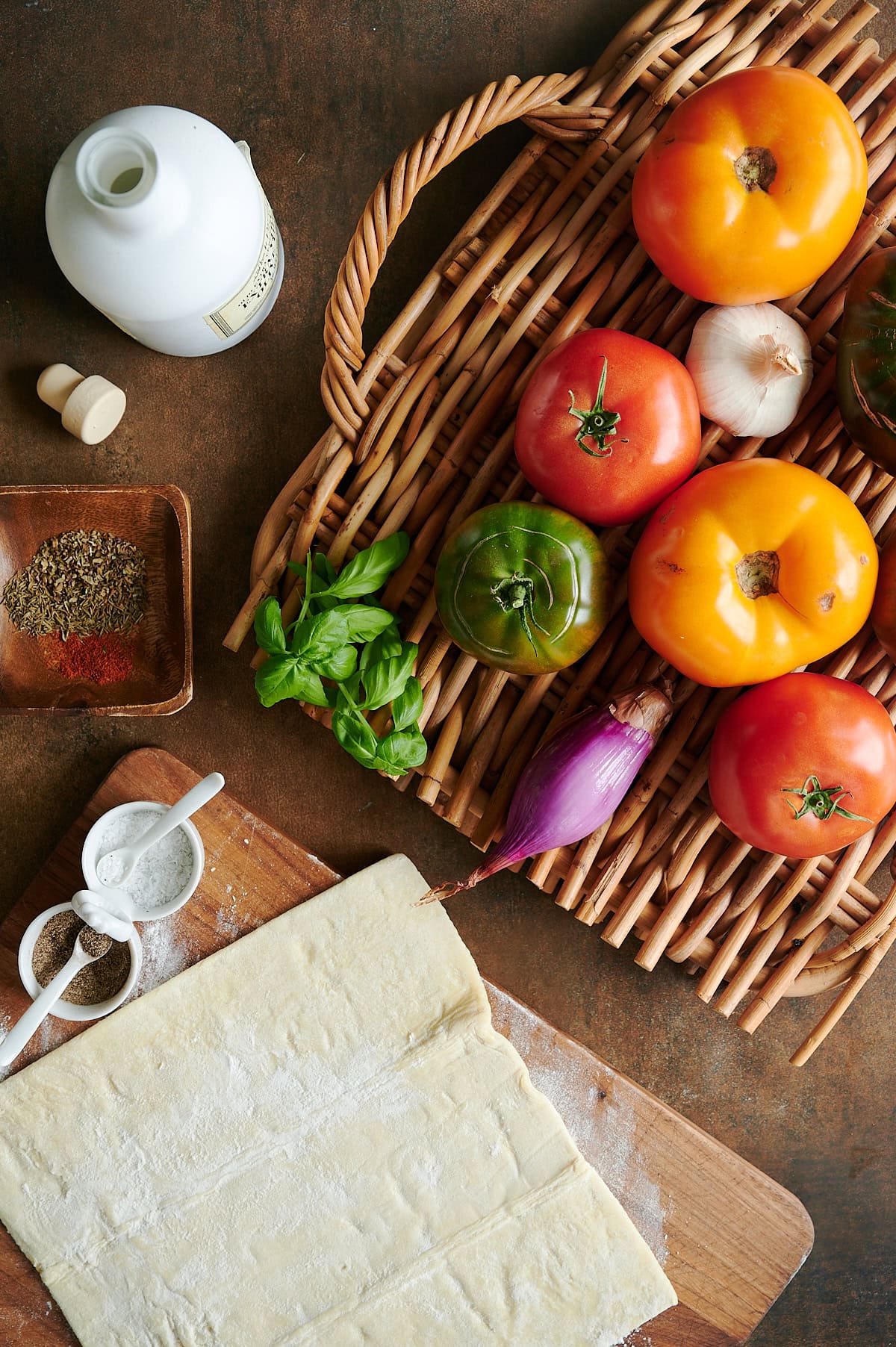 Ingredients for tomato tart on a rattan tray including heirloom tomatoes, puff pastry, olive oil, basil and seasoning.