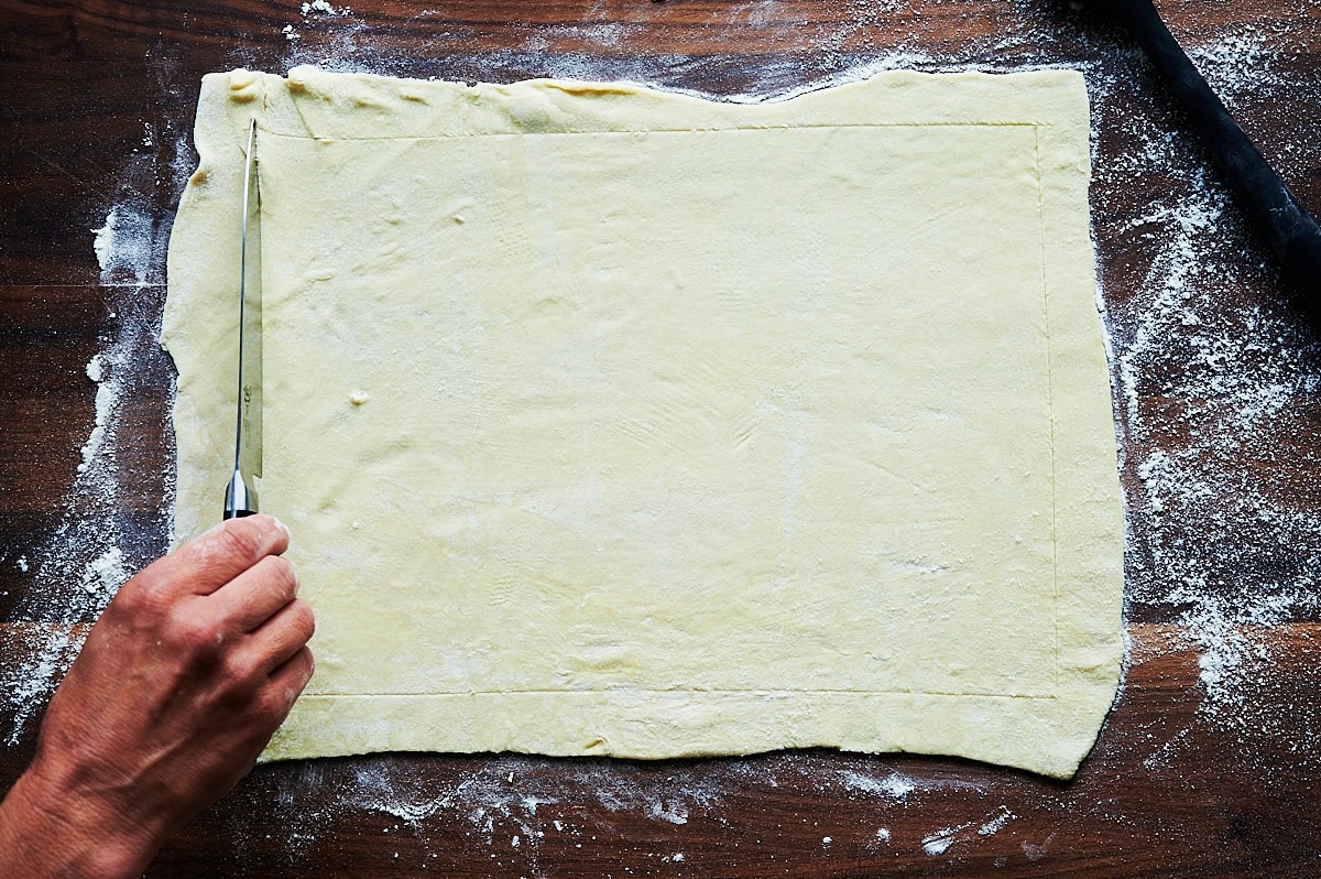 hand scoring a sheet of puff pastry with a knife on a wooden cutting board.
