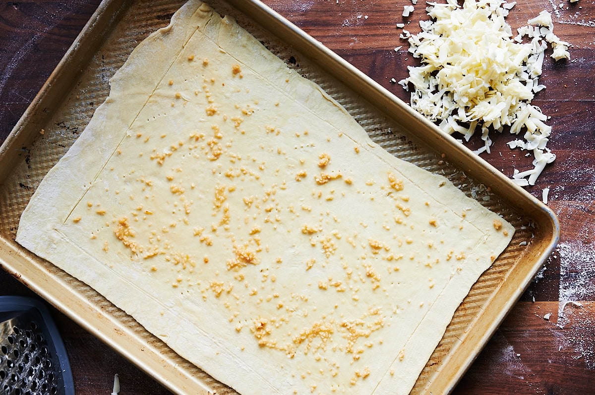 baking sheet on a wooden board containing puff pastry brushed with garlic and shredded cheese on board.