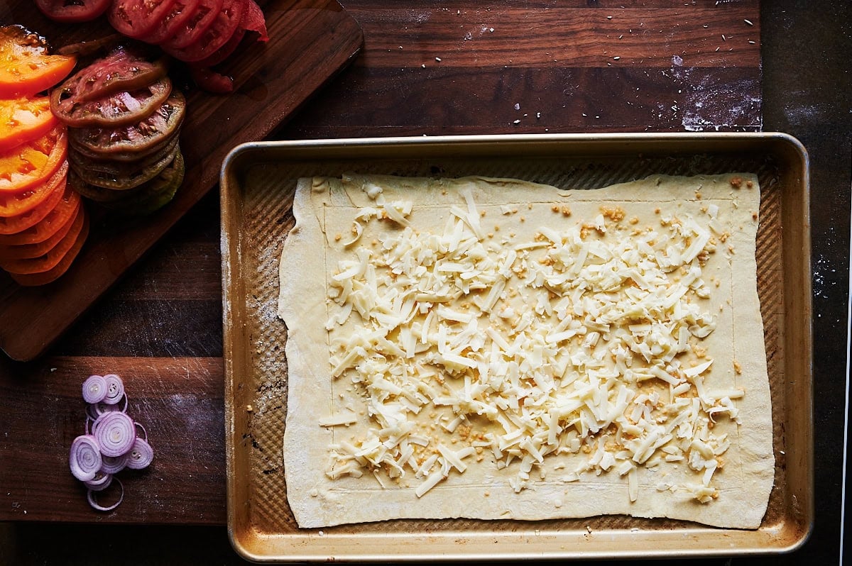 baking sheet with cheese topped puff pastry next to sliced tomatoes and shallots on a wooden board.