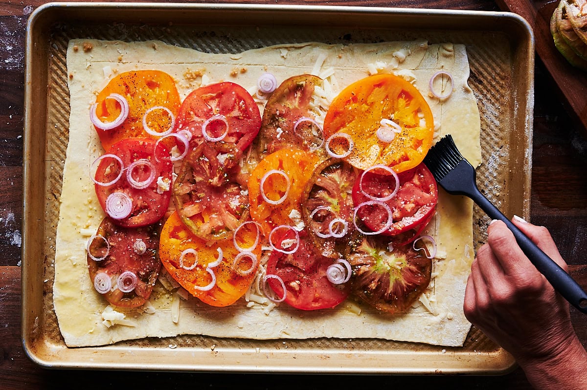 hand brushing egg on edges of a puff pastry tomato tart on a baking sheet.
