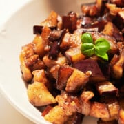 side view of sauteed, basil-garnished, eggplant in a white bowl on a white background.