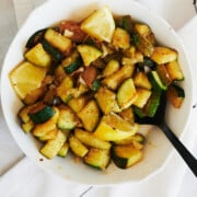 white bowl containing sauteed zucchini with lemon wedges and a black spoon sitting on a white napkin with skinny black lines.