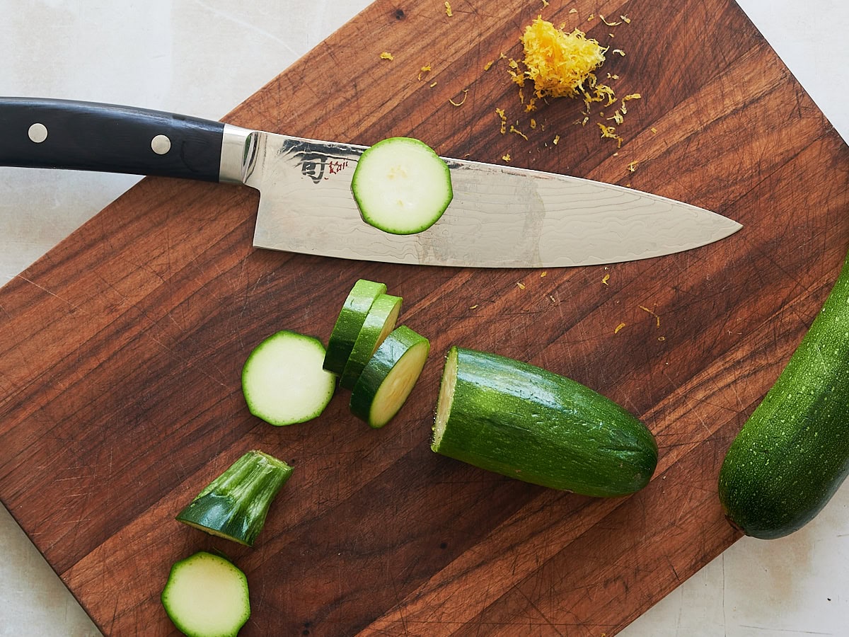 Knife on wooden board slicing zucchini with lemon zest nearby.