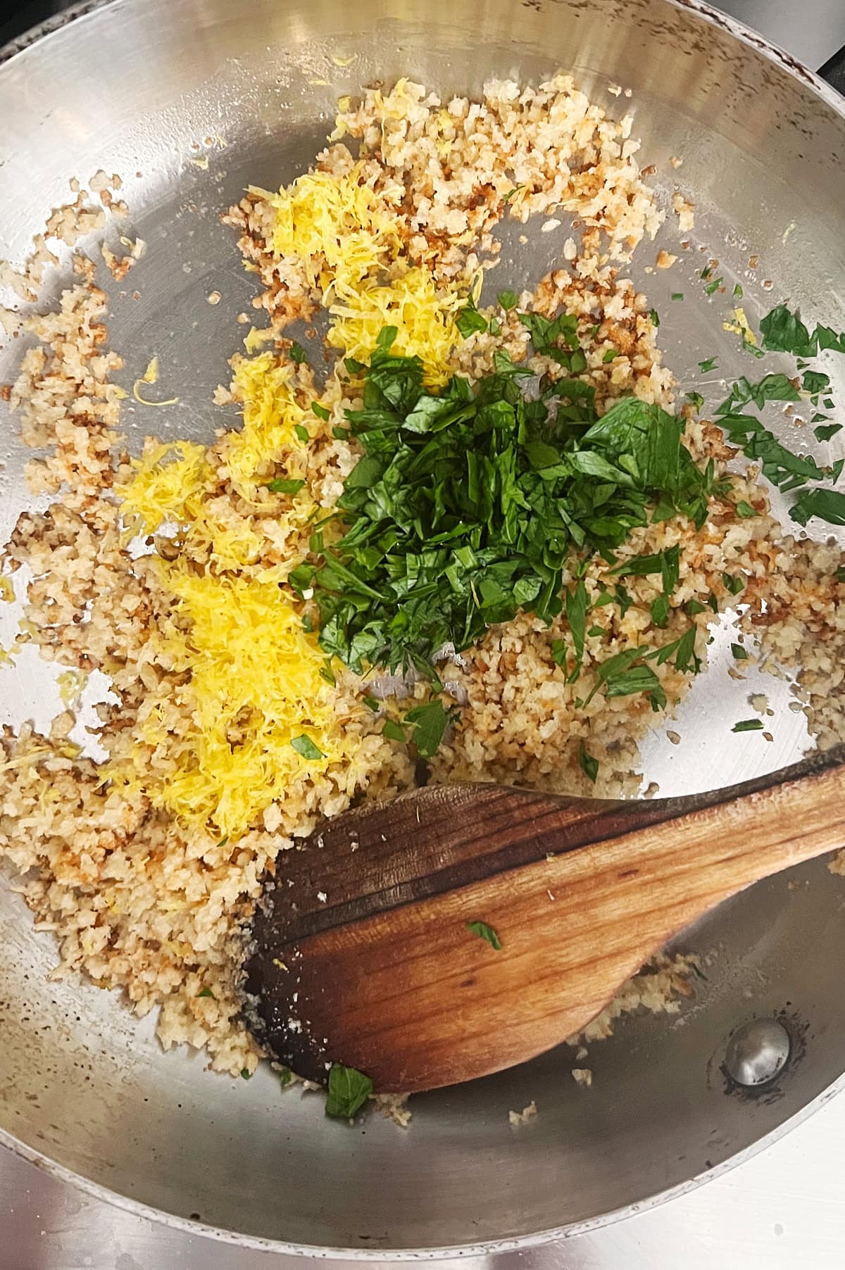 wooden spoon stirring toasted breadcrumbs, lemon zest and parsley in a stainless skillet.