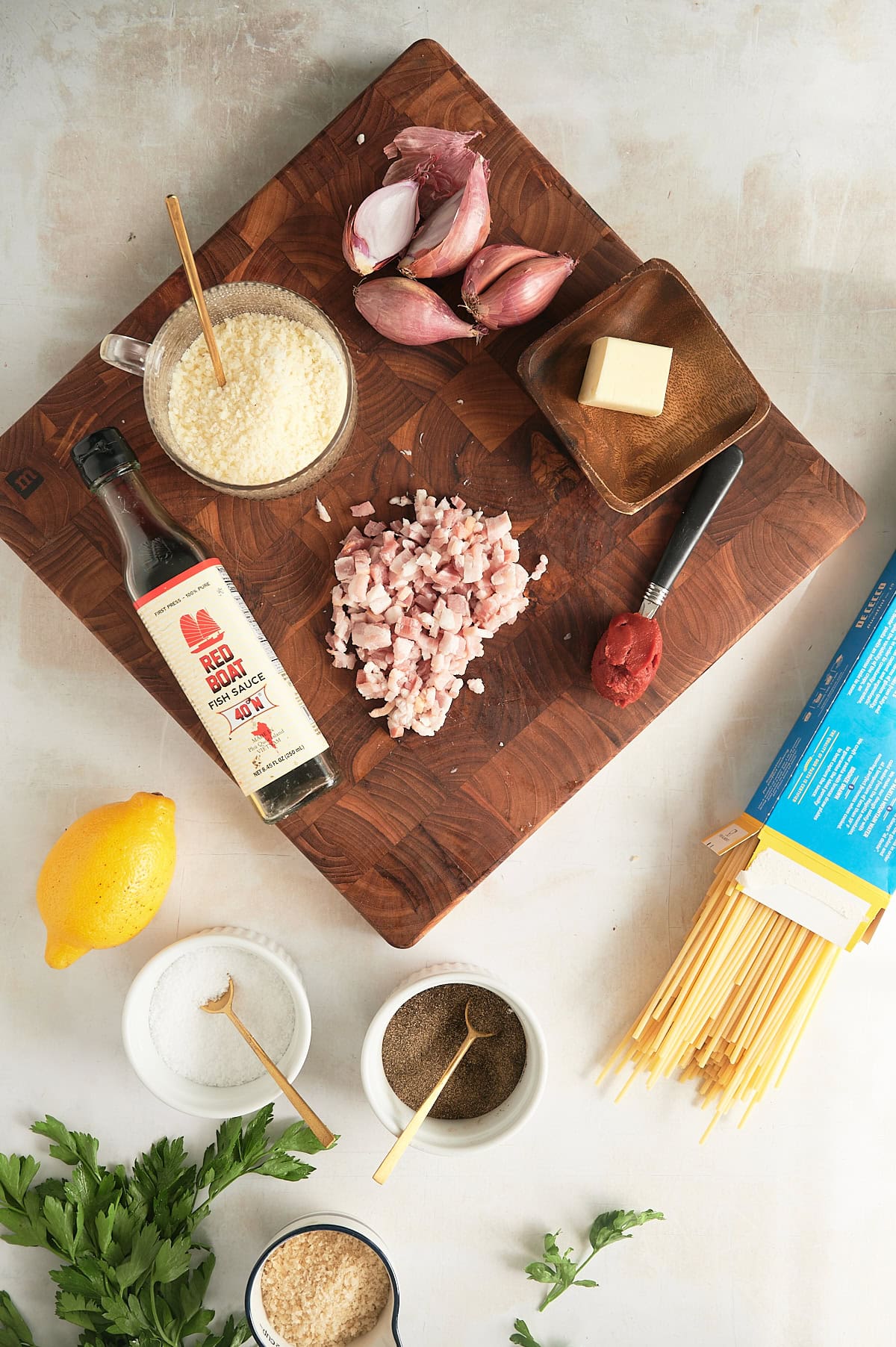 Ingredients for caramelized shallot pasta on wooden board including shallots, tomato paste, lemon, fish sauce, pancetta, bucatini, lemon, breadcrumbs and parsley.