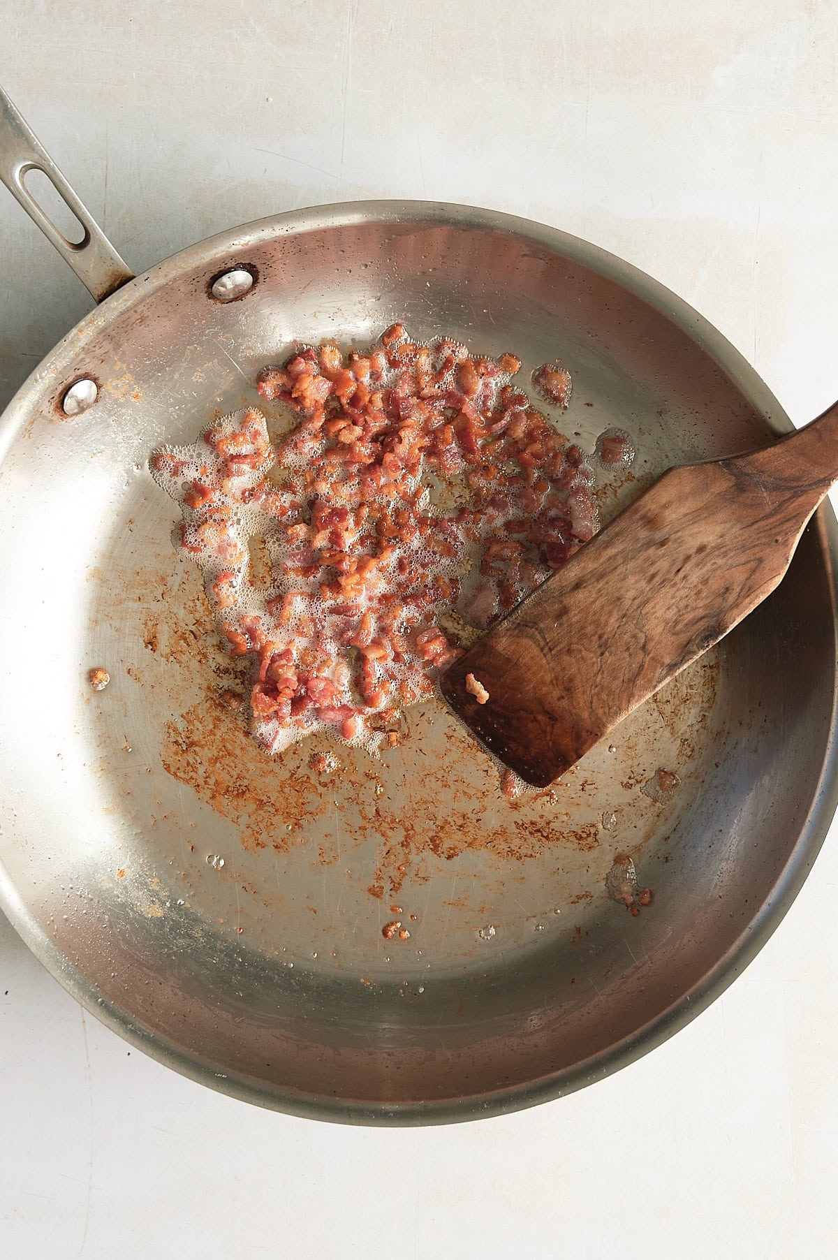 Stainless steel pan of pancetta being sauteed and stirred by a wooden paddle on a cream background.
