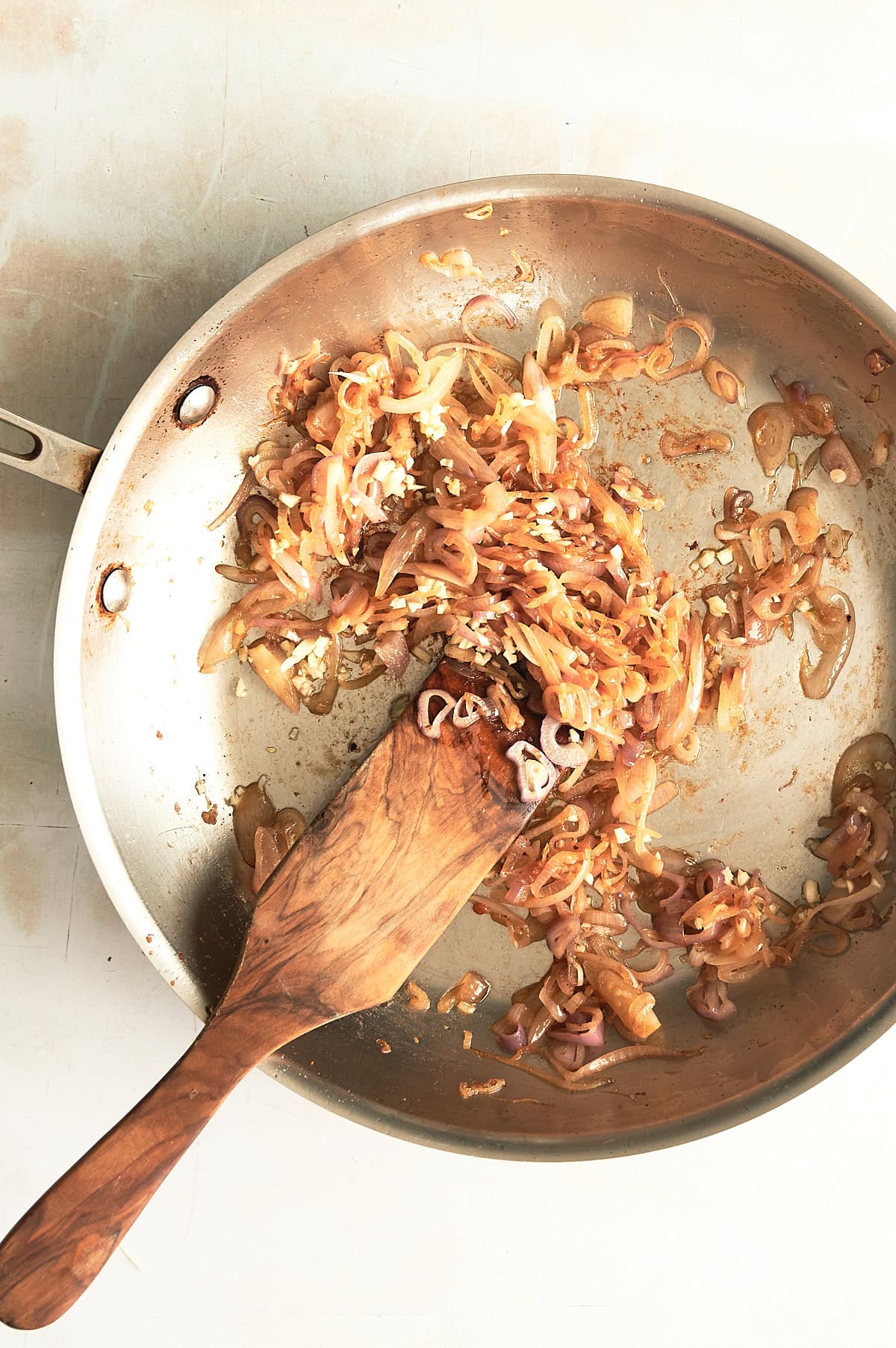 Stainless pan of caramelized shallots with a wooden paddle stirring them on a creamy background.