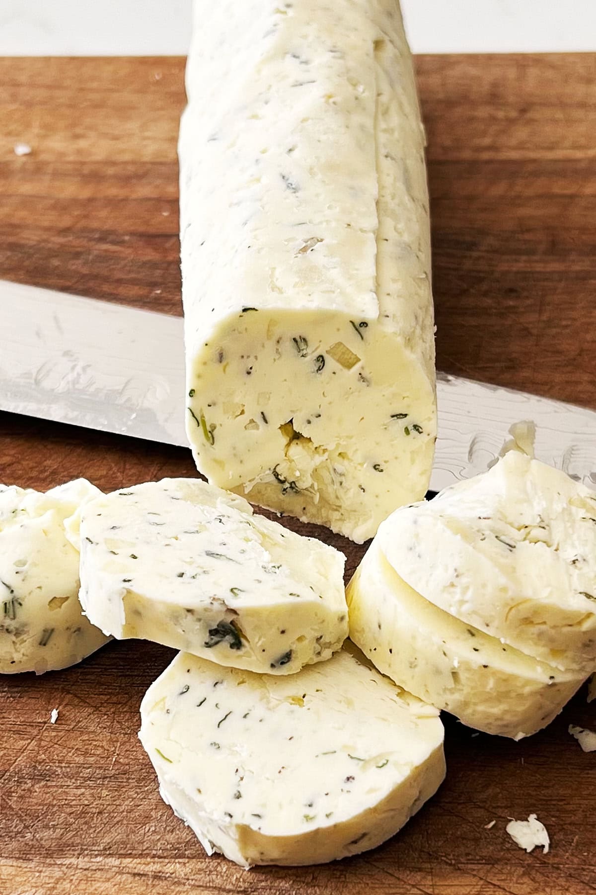 Log of garlic compound butter on a wooden board being sliced with a knife.