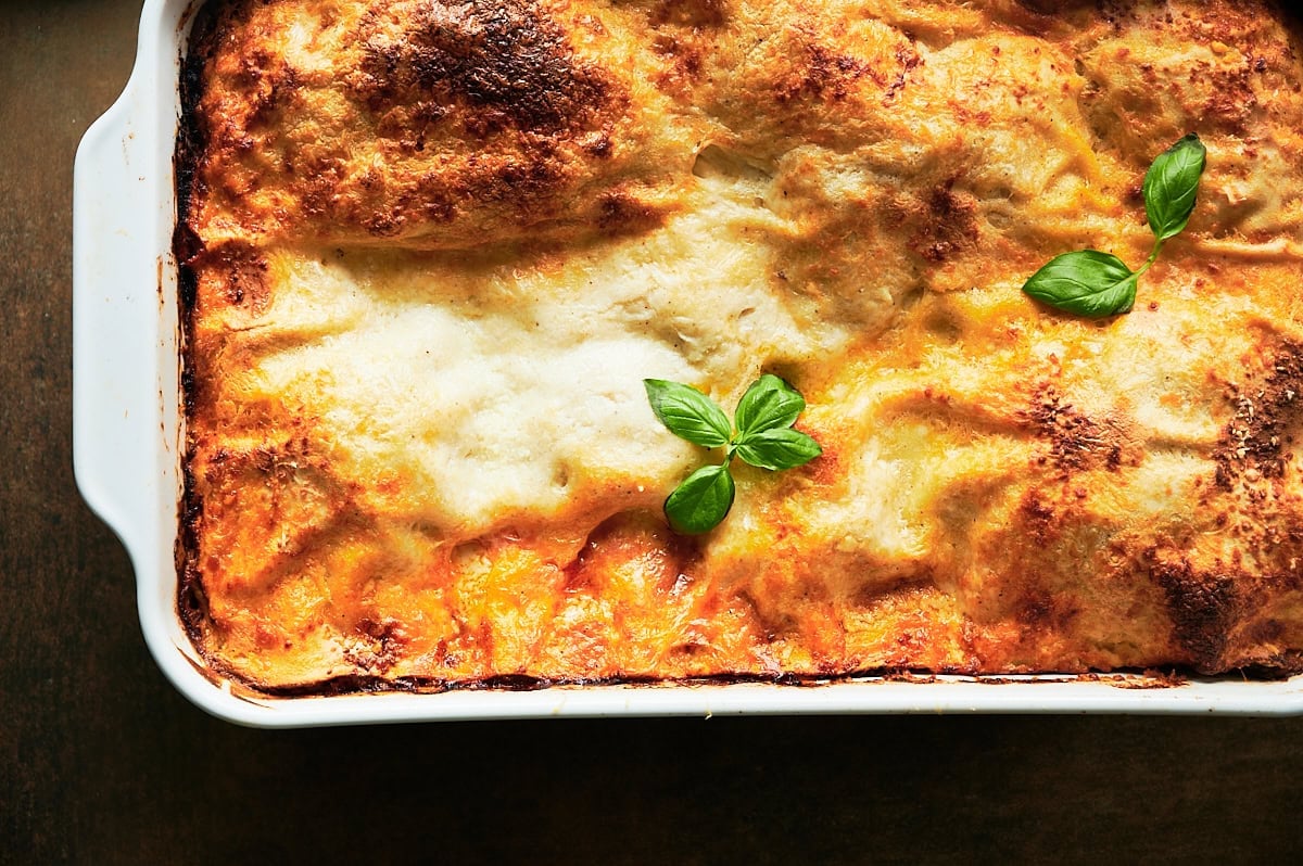 close up of a pan of crispy baked pan lasagna bolognese in a white baking dish with fresh basil leaves on top.