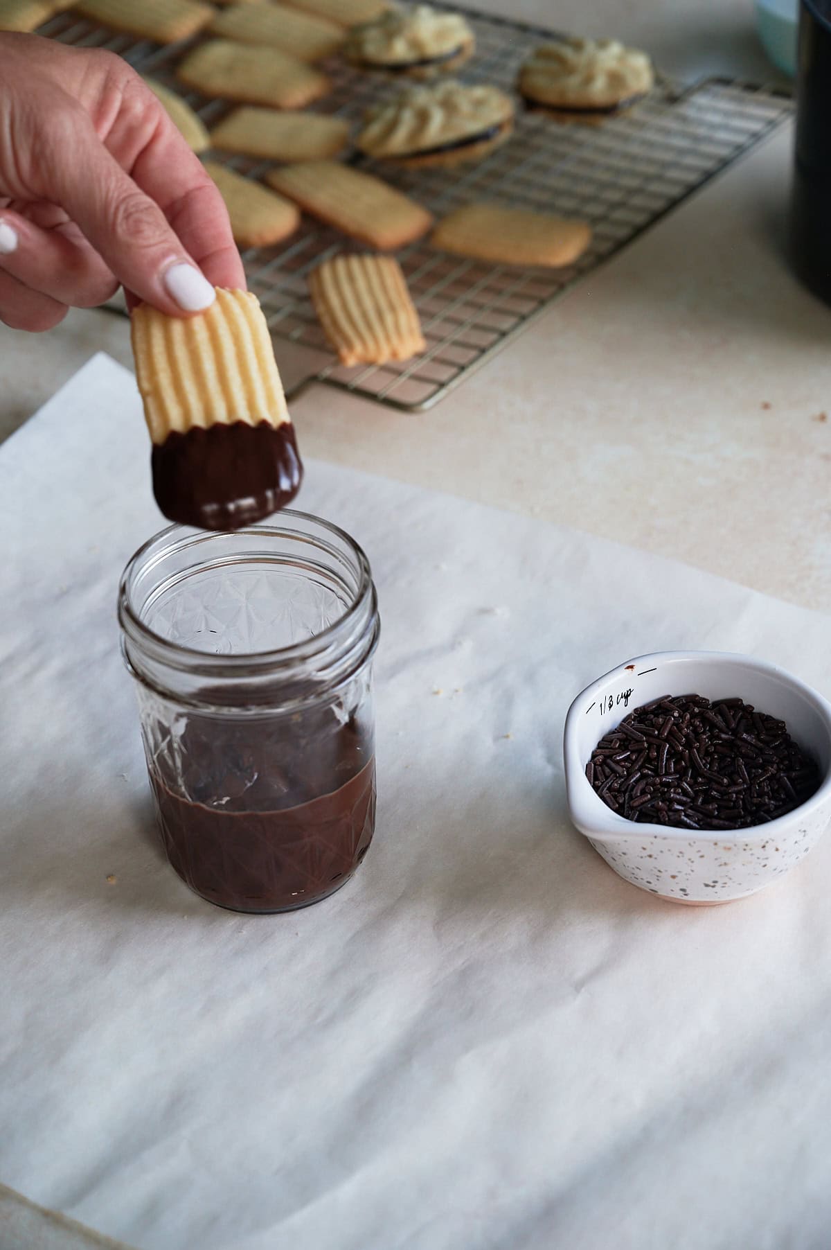 Hand dipping end of a butter cookie in a small jelly glass of chocolate ganache with a small sprinkle bowl of chocolate jimmies.