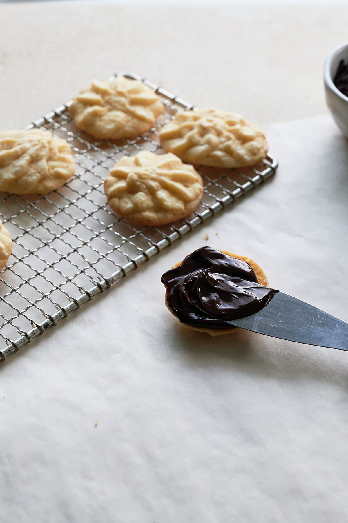 Spatula spreading chocolate ganache on one half of butter cookie next to cooling rack of star butter cookies.