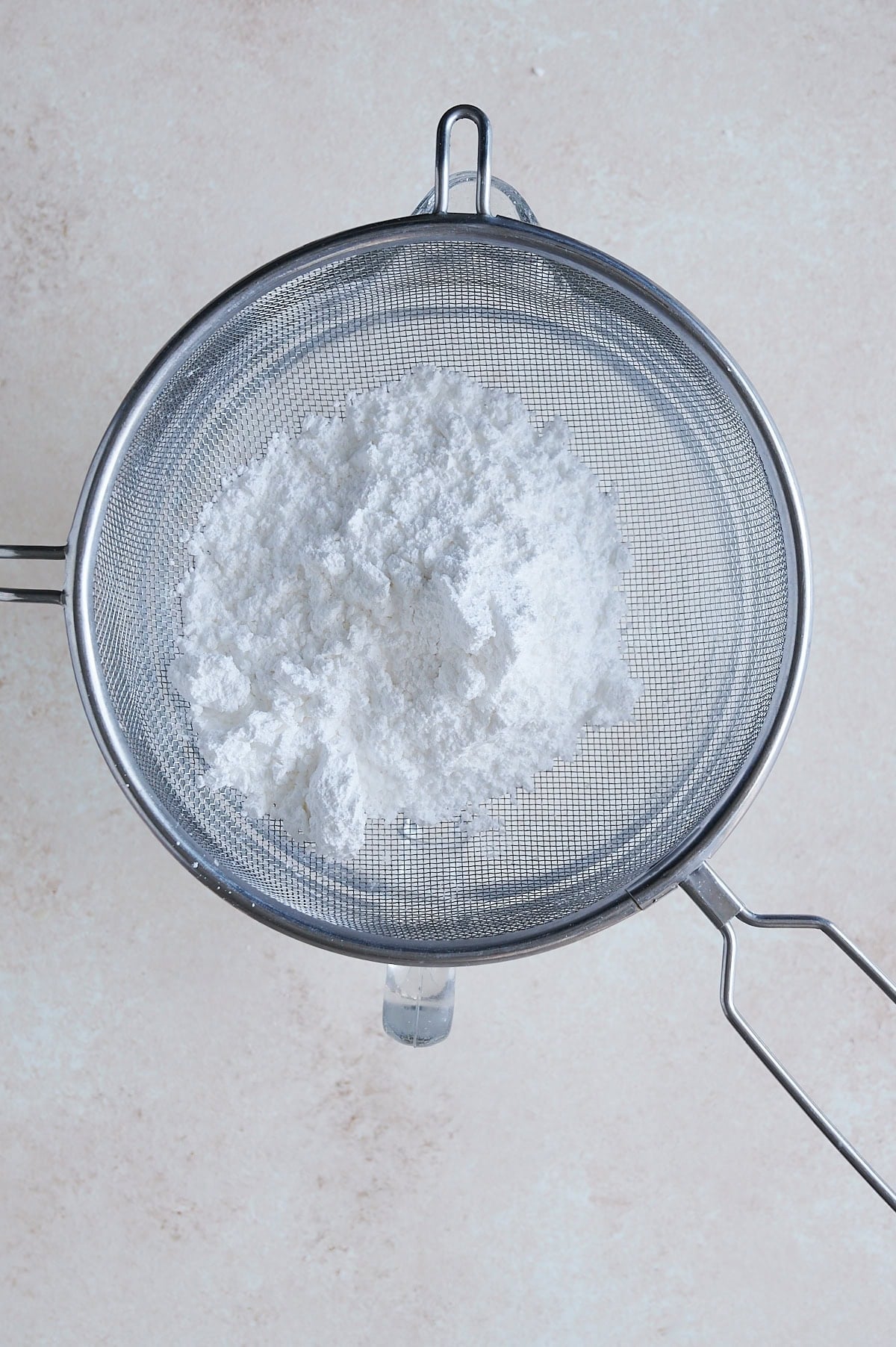 A wire mesh strainer with confectioners sugar in it over a bowl on a cream background.