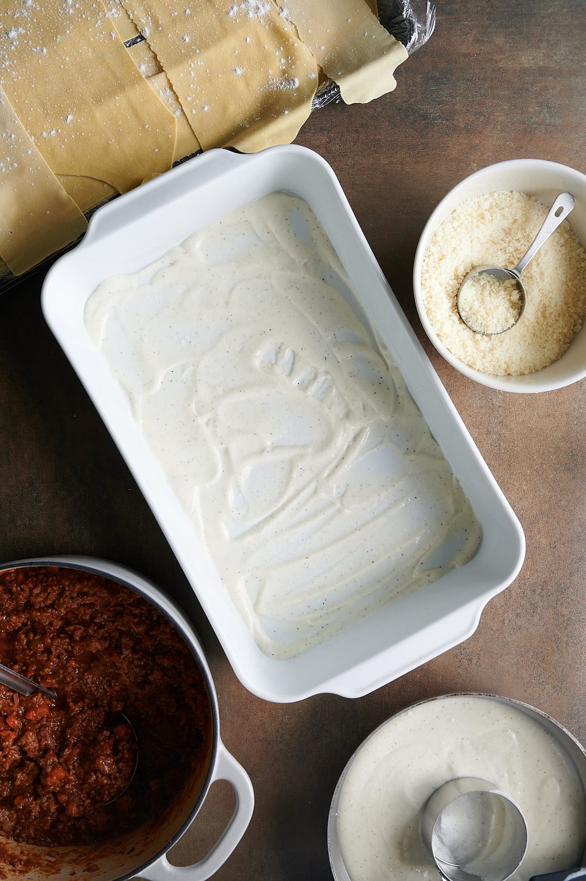 Bechamel sauce spread all over the bottom of a baking dish for lasagna Bolognese with bowls and pan of lasagna noodles, Bolognese sauce and grated parmesan cheese.