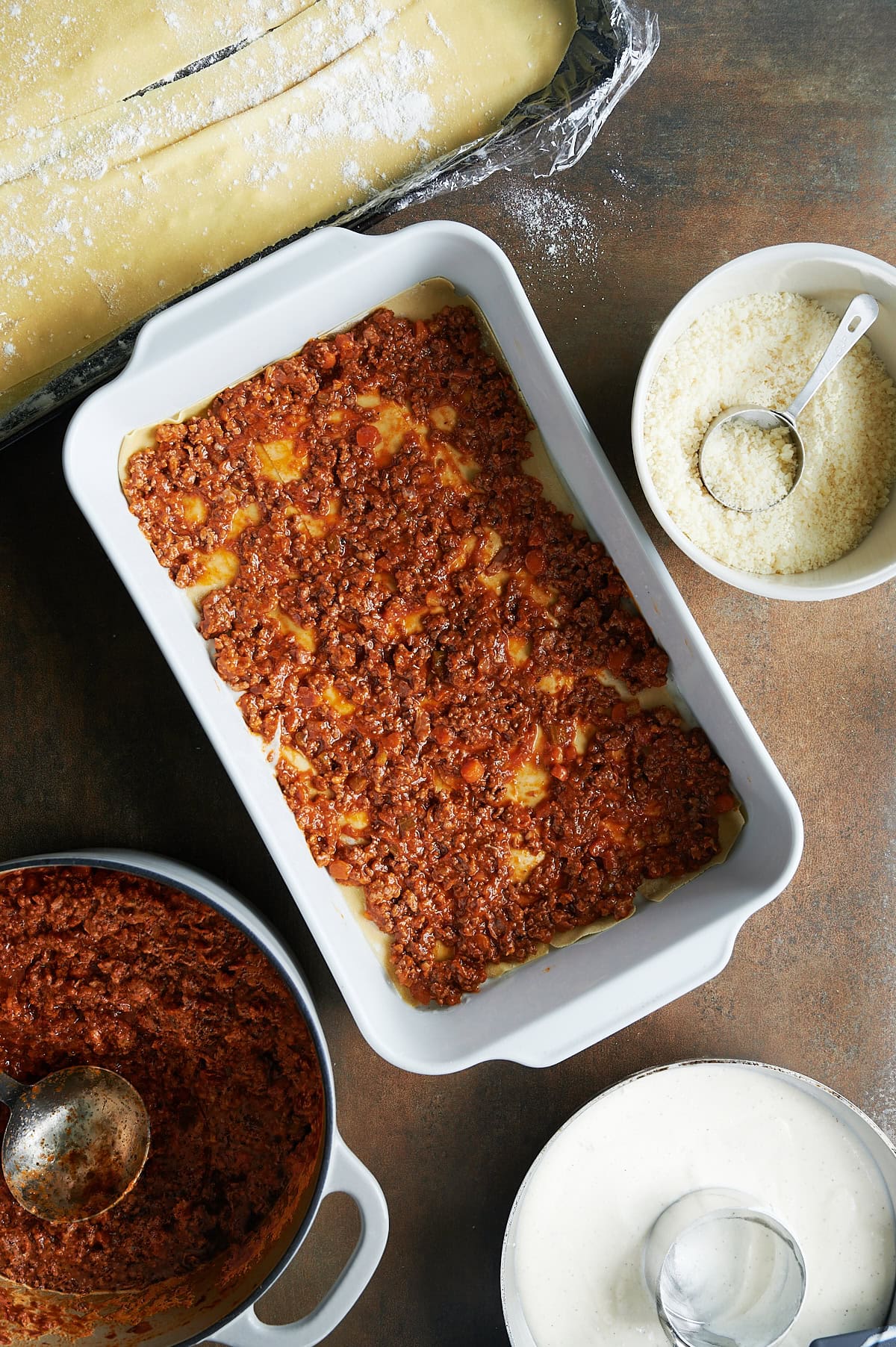 White baking dish with layer 1 of Bolognese sauce layered on lasagna noodles with bowls of parmesan, bechamel, Bolognese sauce nearby.