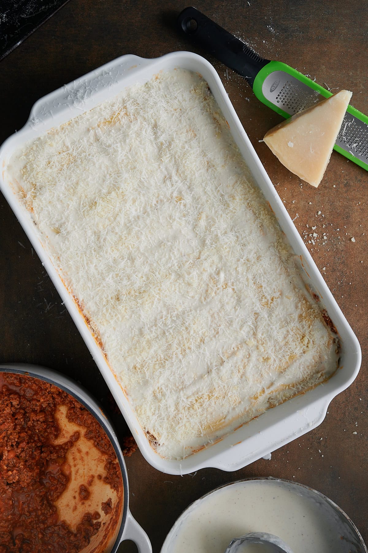 Unbaked pan of lasagna Bolognese with Bolognese sauce, bechamel and a wedge of parmesan cheese on a grater.