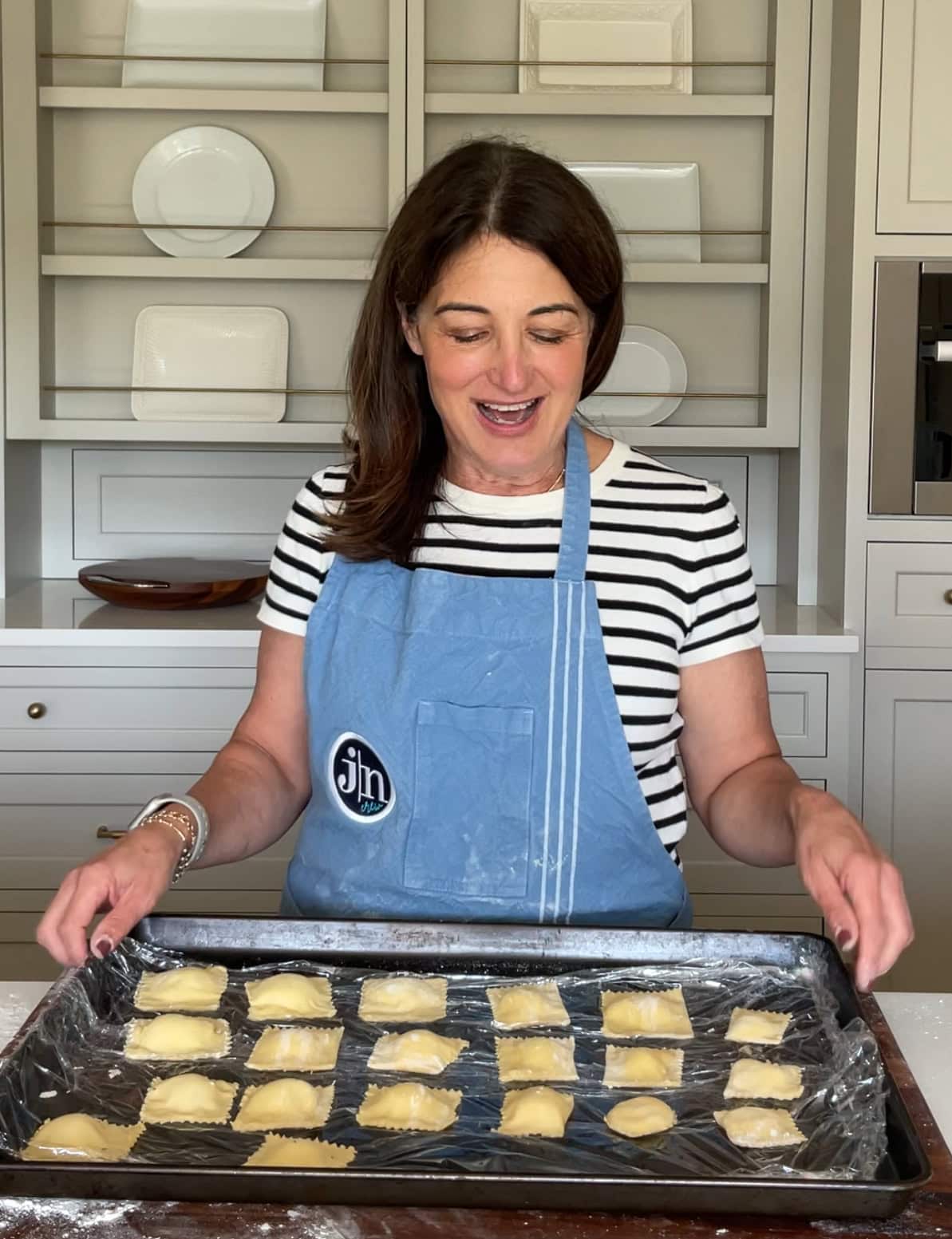 brunette woman in a blue apron and black and cream stripe shirt making ravioli on a wooden cutting board.