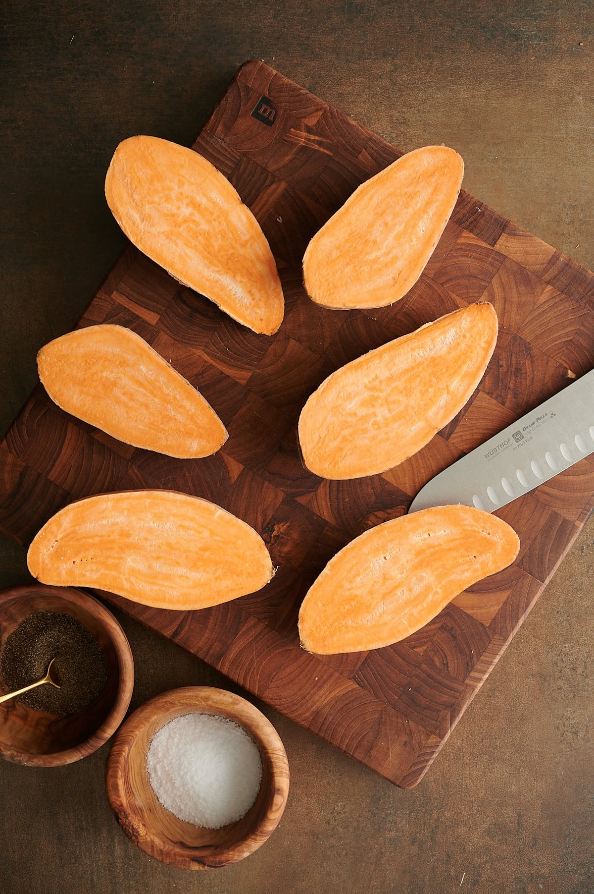 Three cut sweet potato halves arranged on a wooden board with bowls of salt and black pepper.