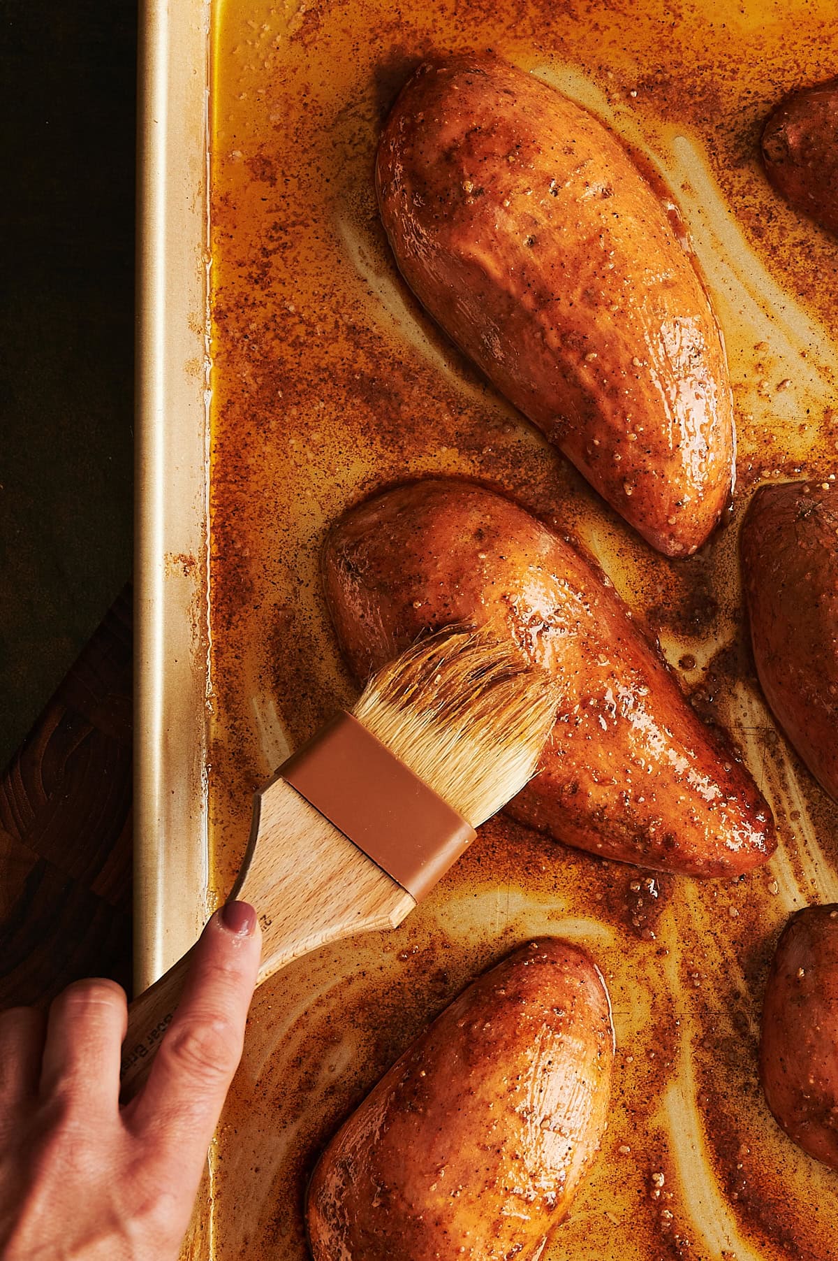 Hand brushing seasoned butter over tops of sweet potatoes on a gold sheet pan.