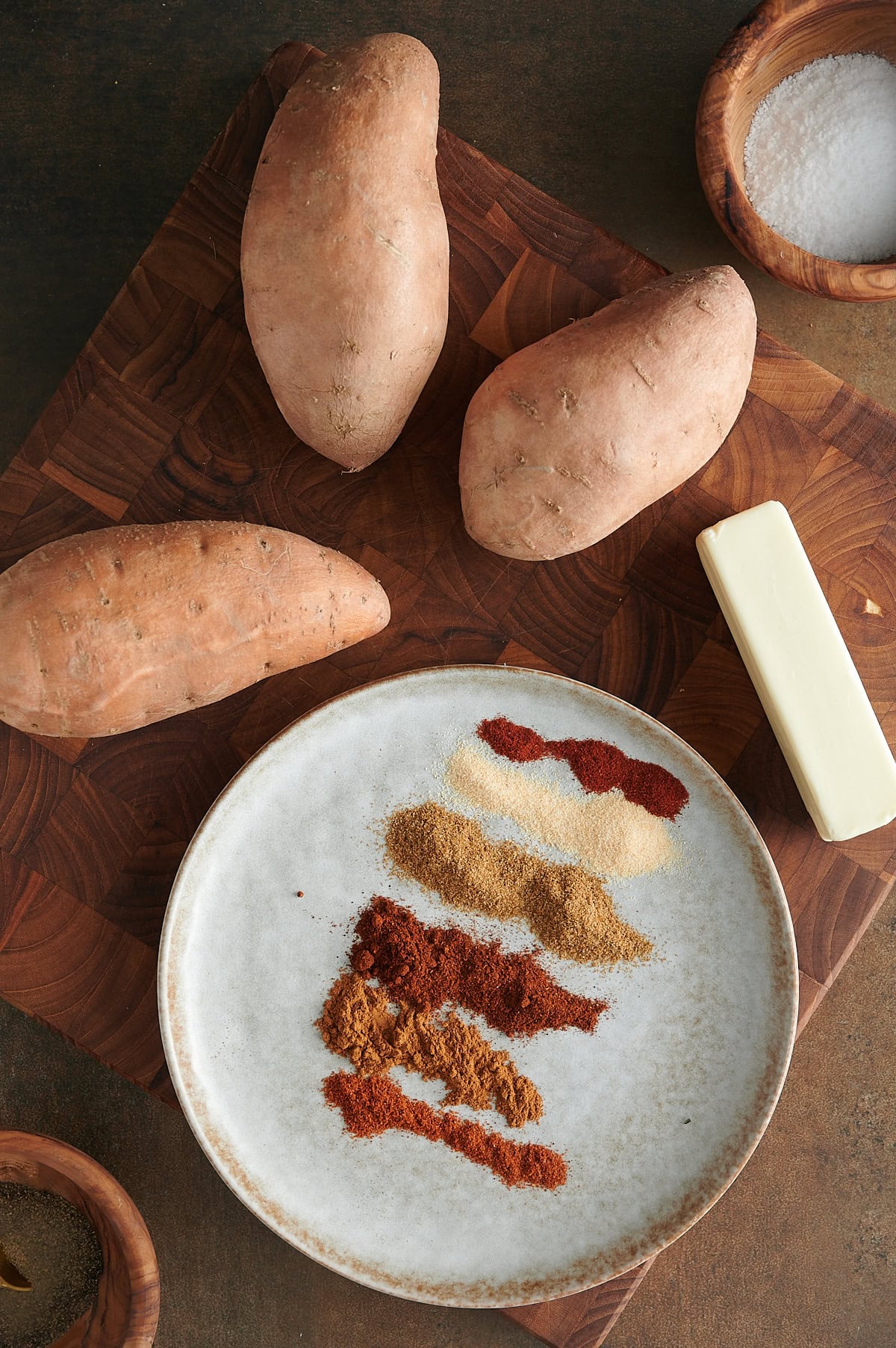 Ingredients for roasted sweet potatoes on a white plate with potatoes and stick of butter on a cutting board.