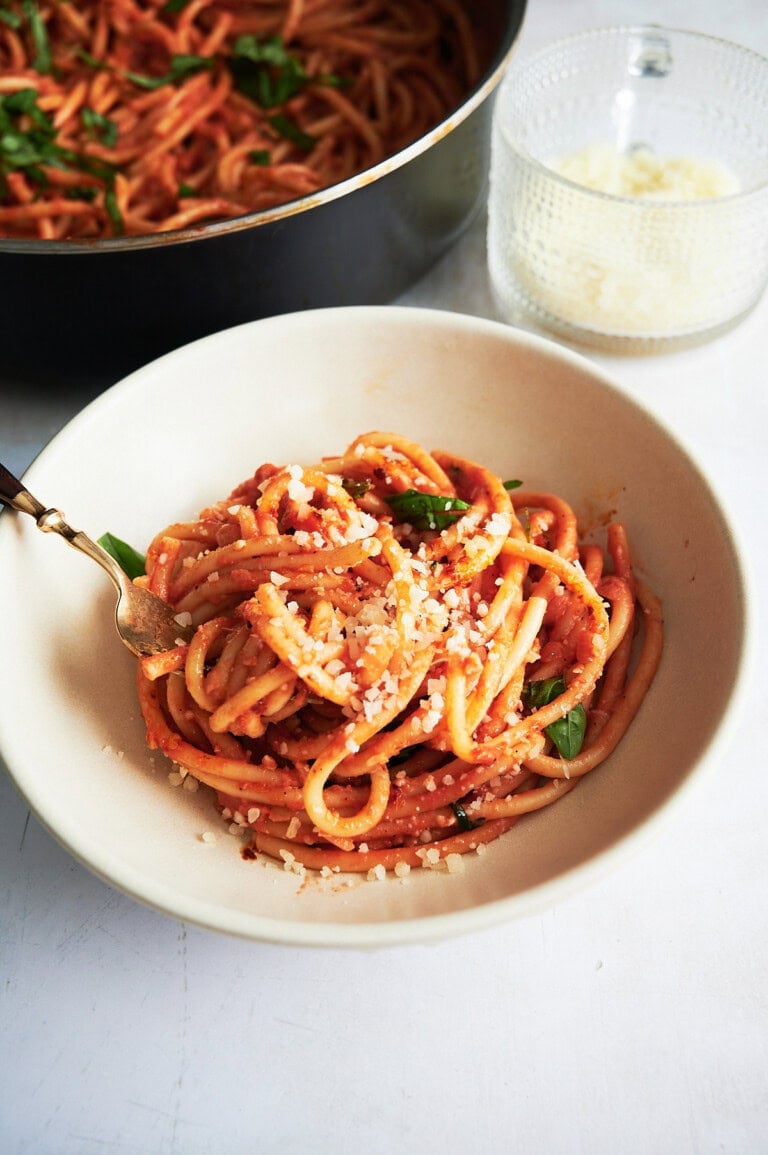 White bowl of bucatini with tomato mascarpone sauce sprinkled with fresh basil and parmesan cheese.