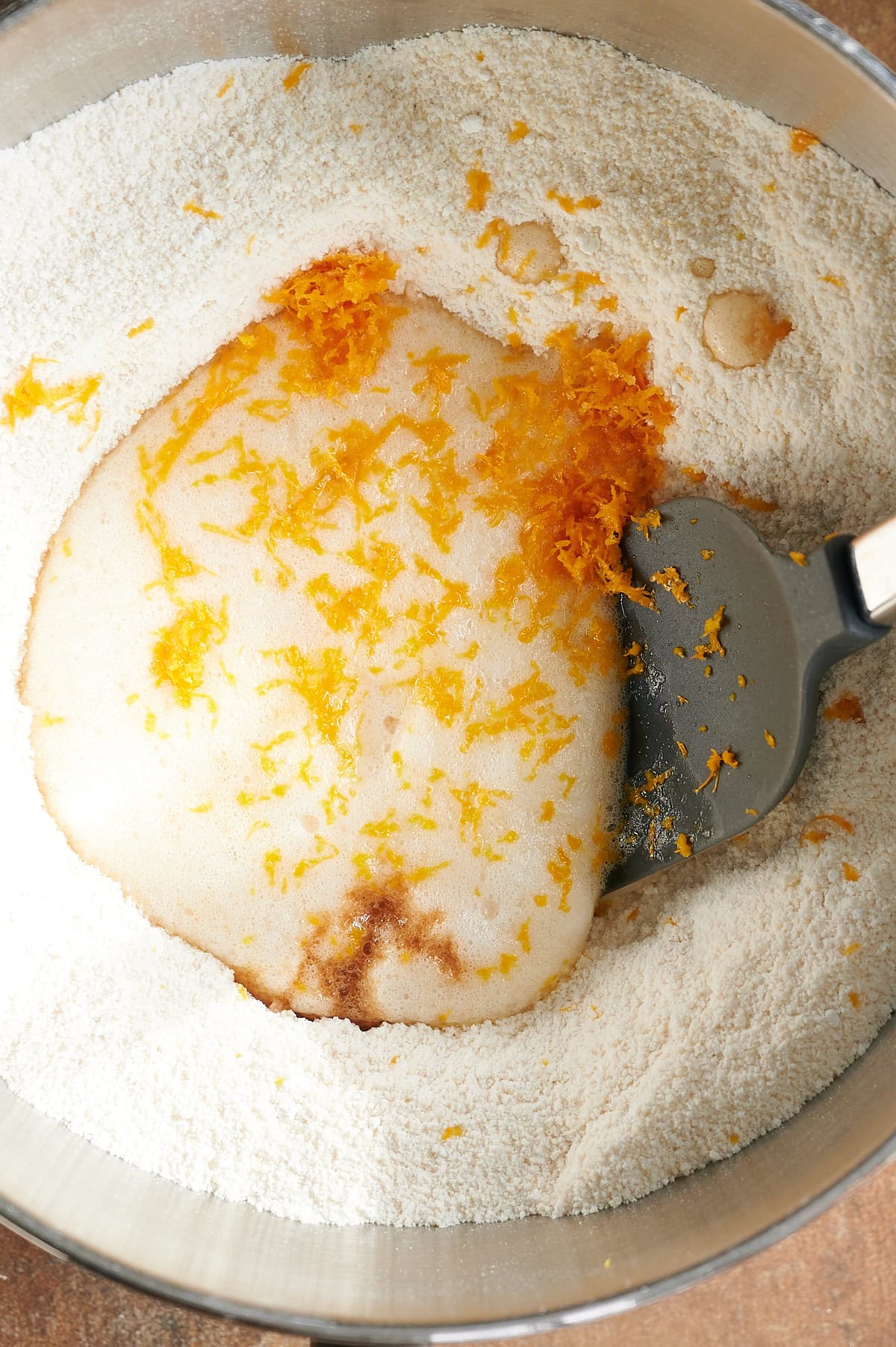 Gray spatula folding orange zest and egg whites into dry ingredients for almond cookies.