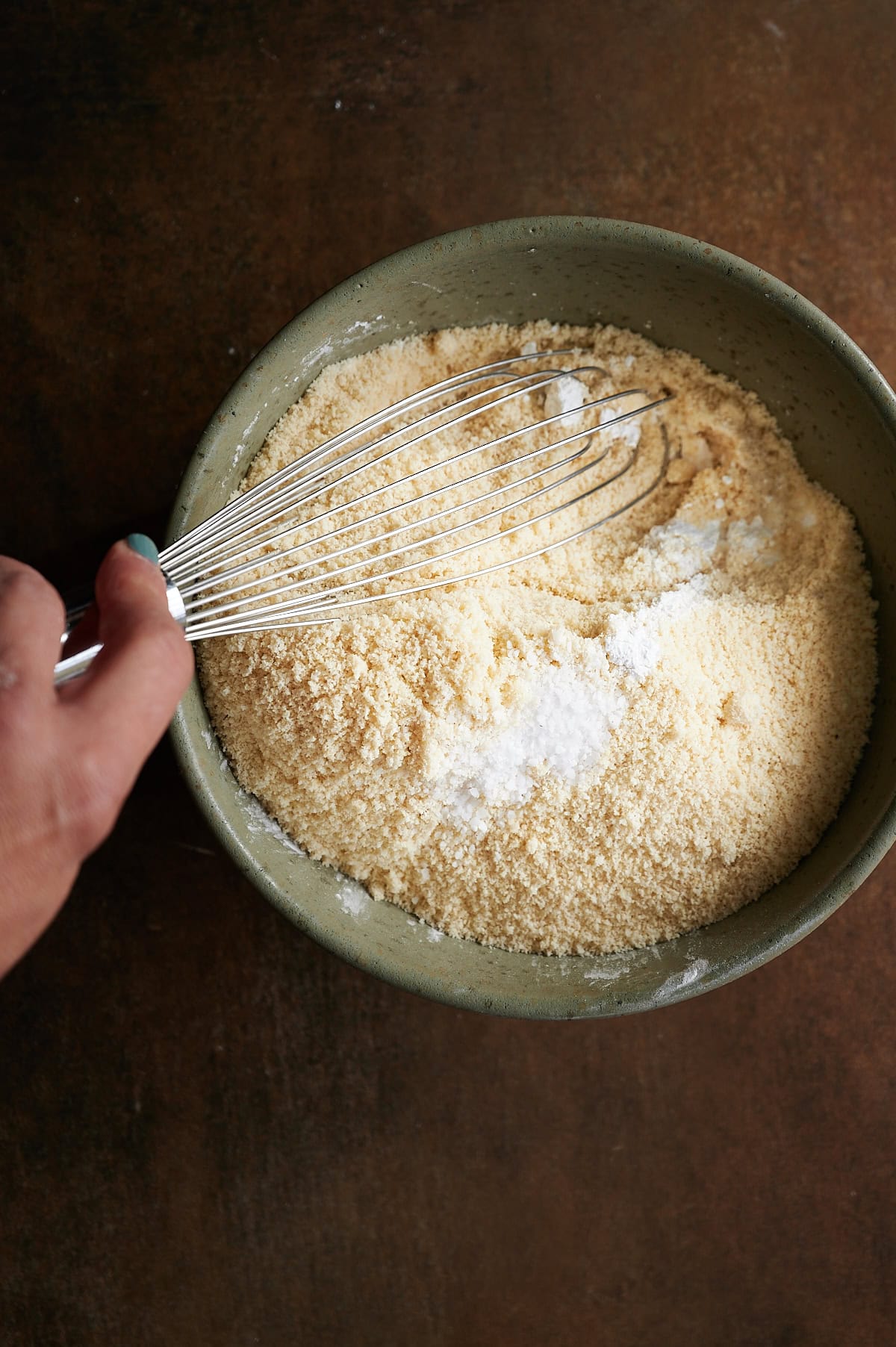 Green bowl on a brown background with almond flour and confectioners sugar being whisked.