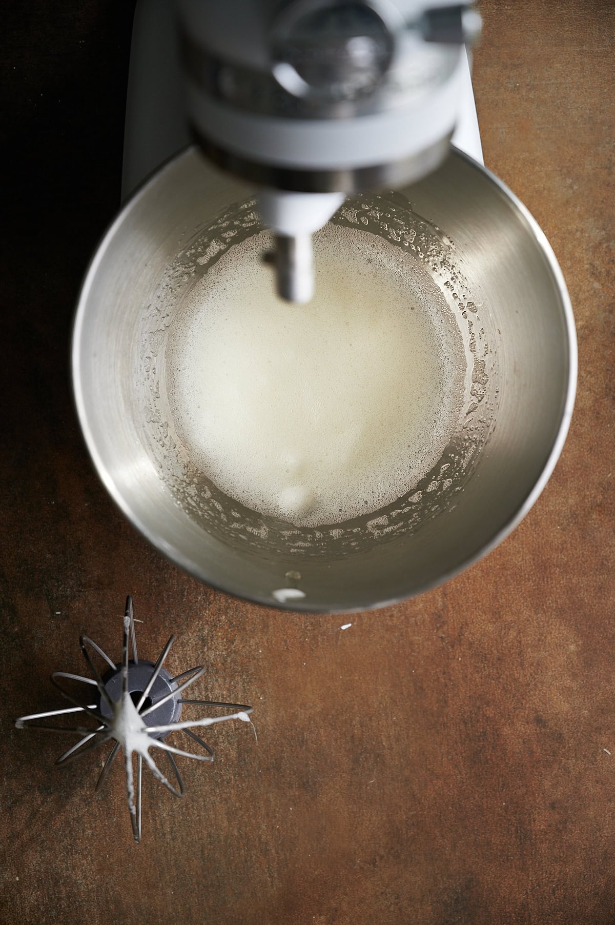 Whipped egg white in stainless mixing bowl on brown background with stainless whisk beater near by.