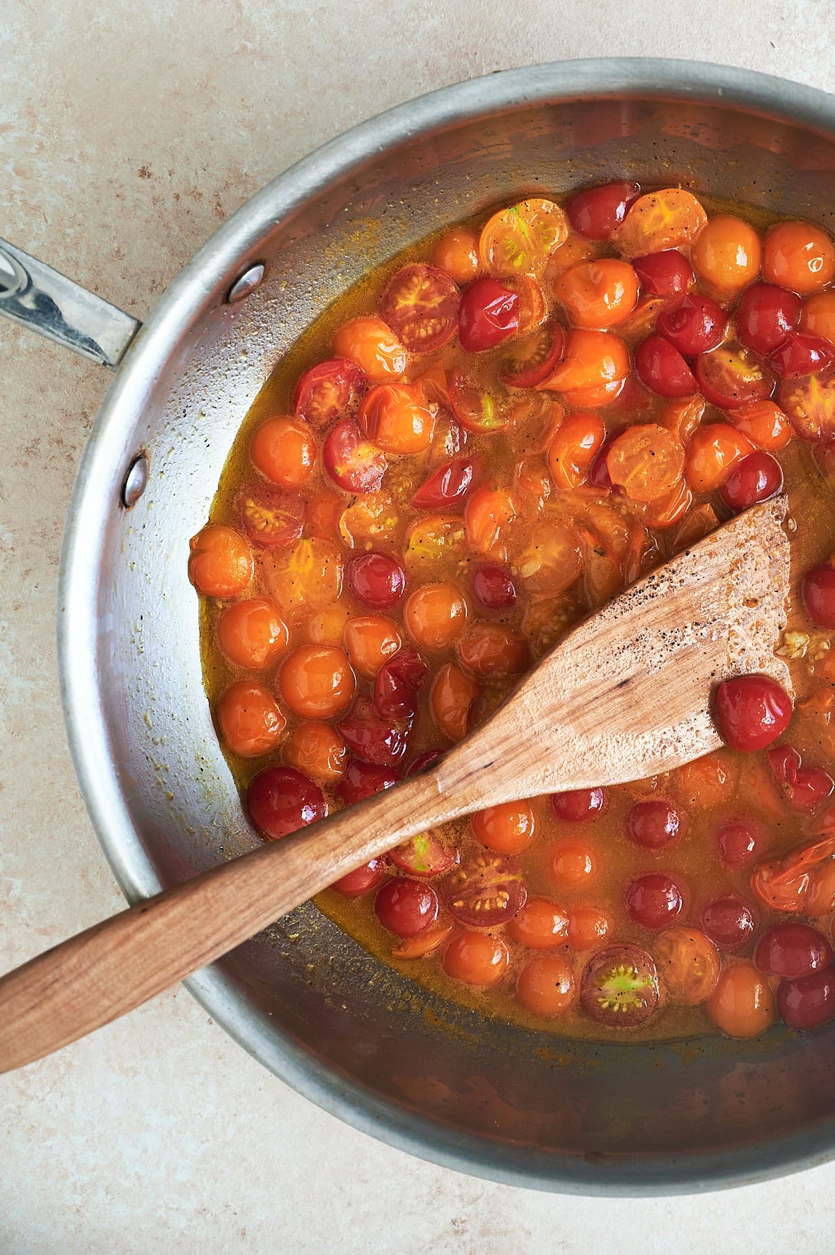 Stainless skillet of red and orange cherry tomato sauce with garlic being cooked with wooden paddle.