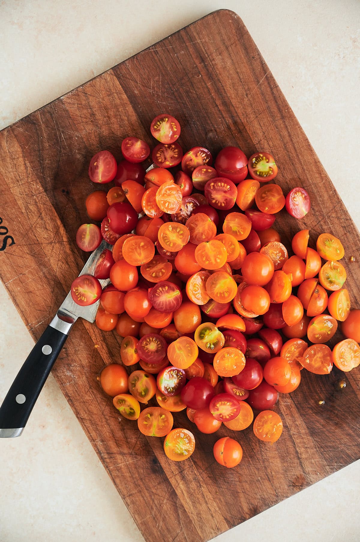 Knife with tomatoes being cut in half on wooden cutting board.