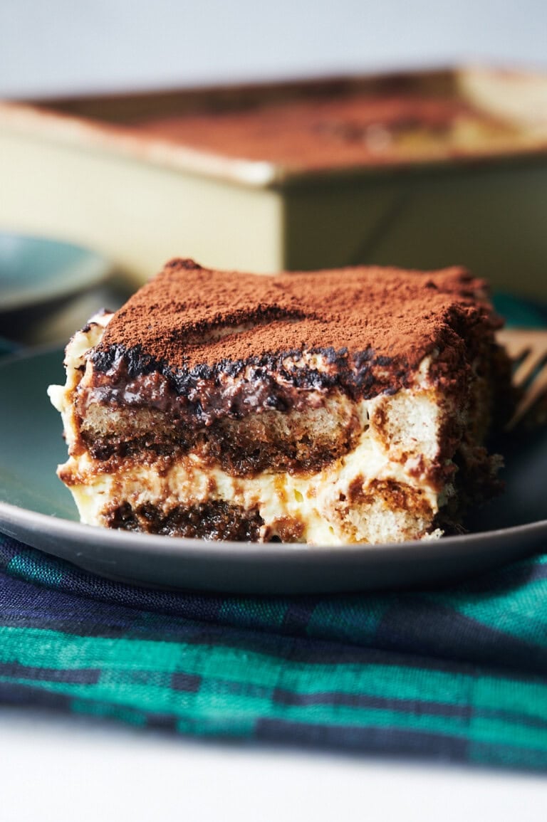 a piece of cocoa sprinkled tiramisu on a black plate and a blackwatch napkin.