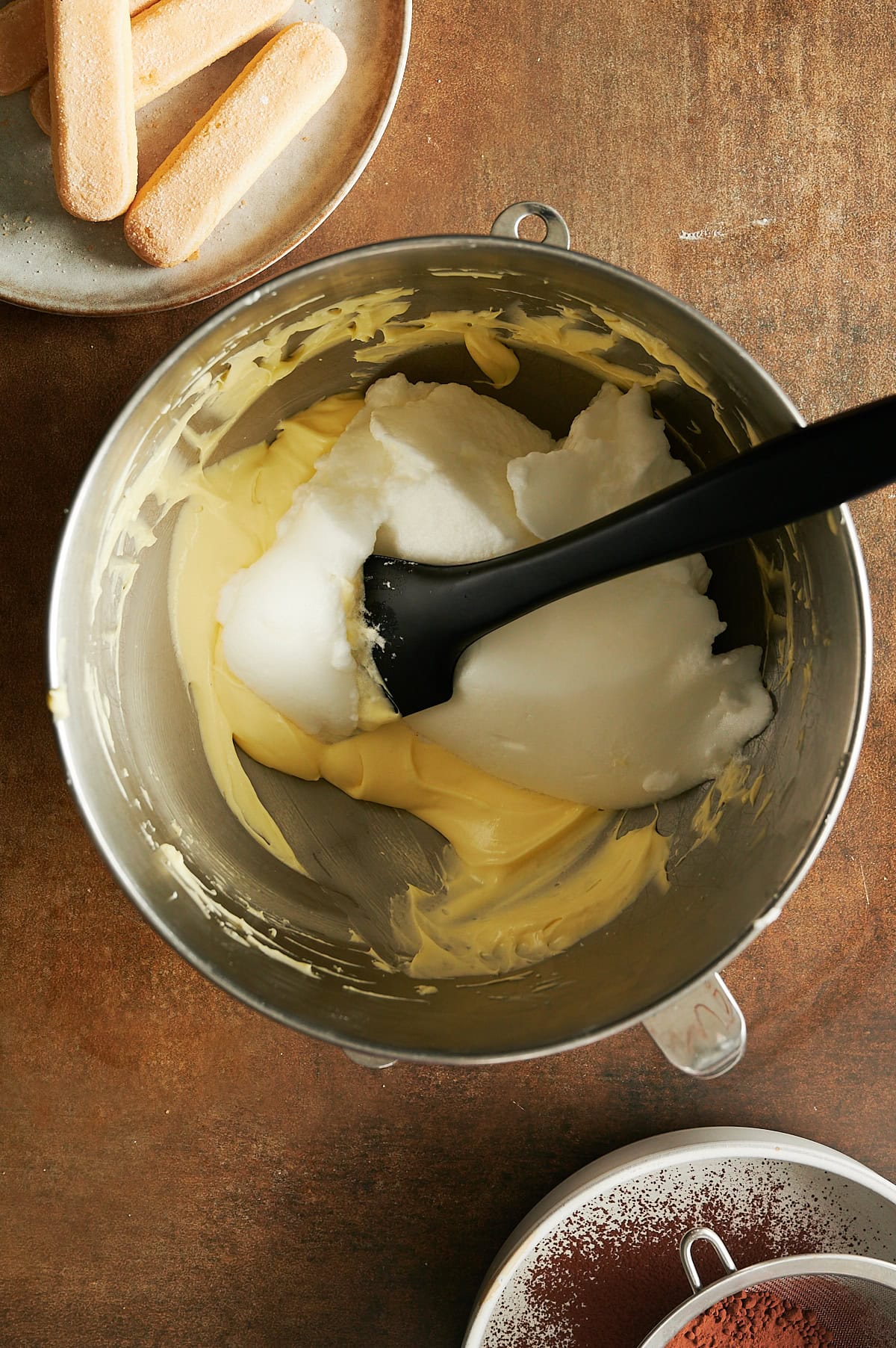 Mixing bowl of whipped egg whites being folded into egg yolk cream for tiramisu with lady fingers and cocoa powder nearby.