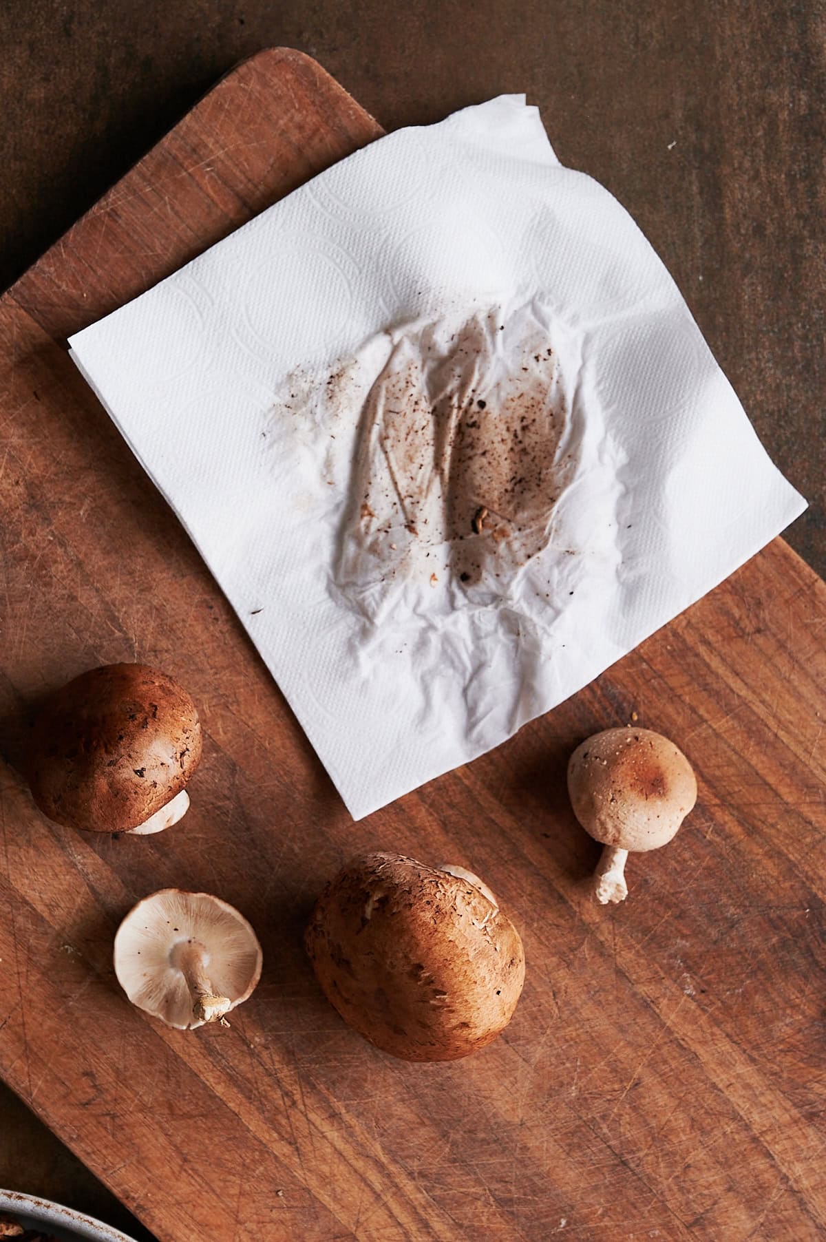 Brown cutting board with mushrooms being cleaned by a white towel all on a brown background.