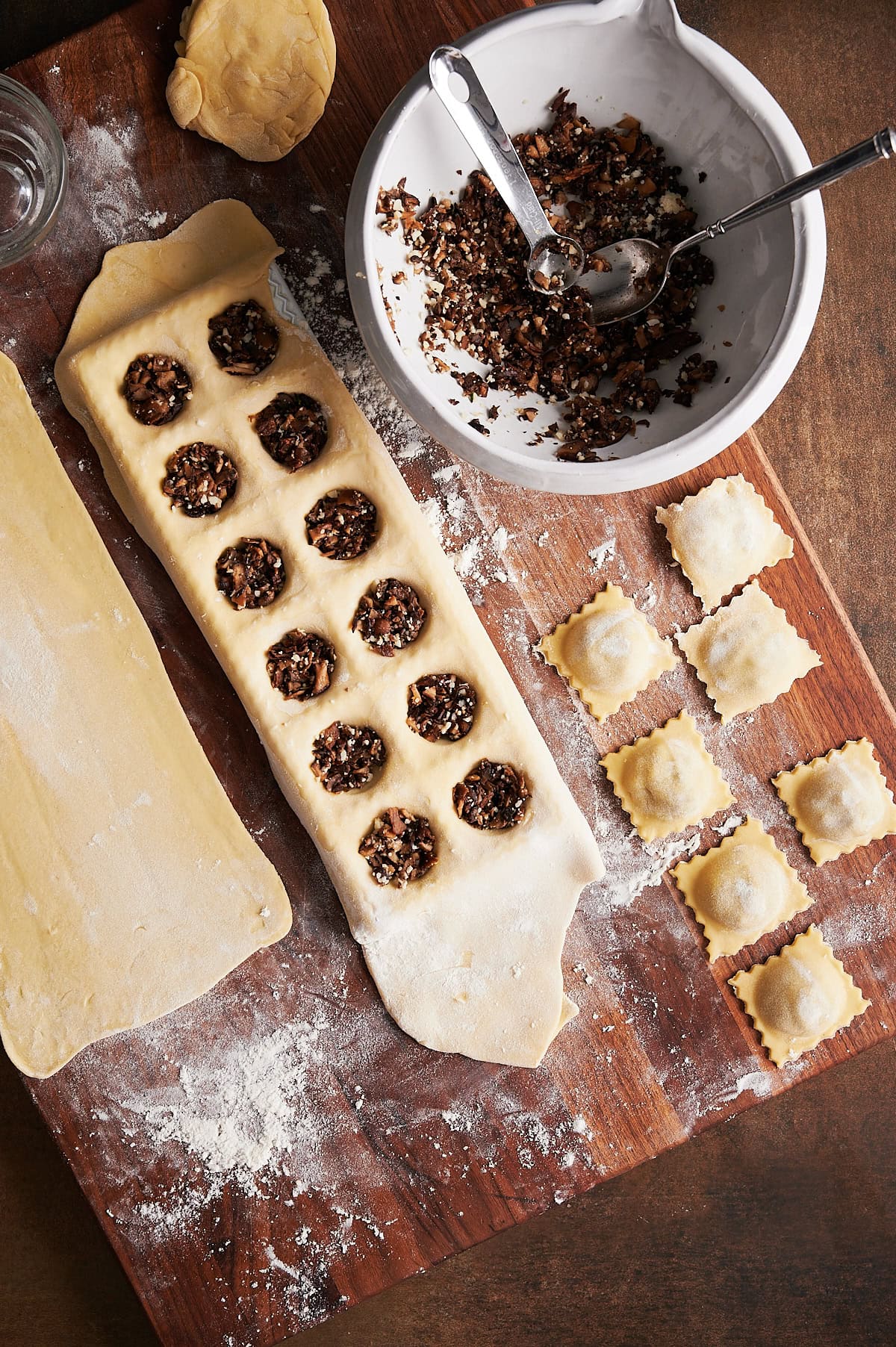 Brown board holding rolled ravioli dough sheet with a ravioli mold filled with mushroom filling and bowl of filling and finished ravioli.