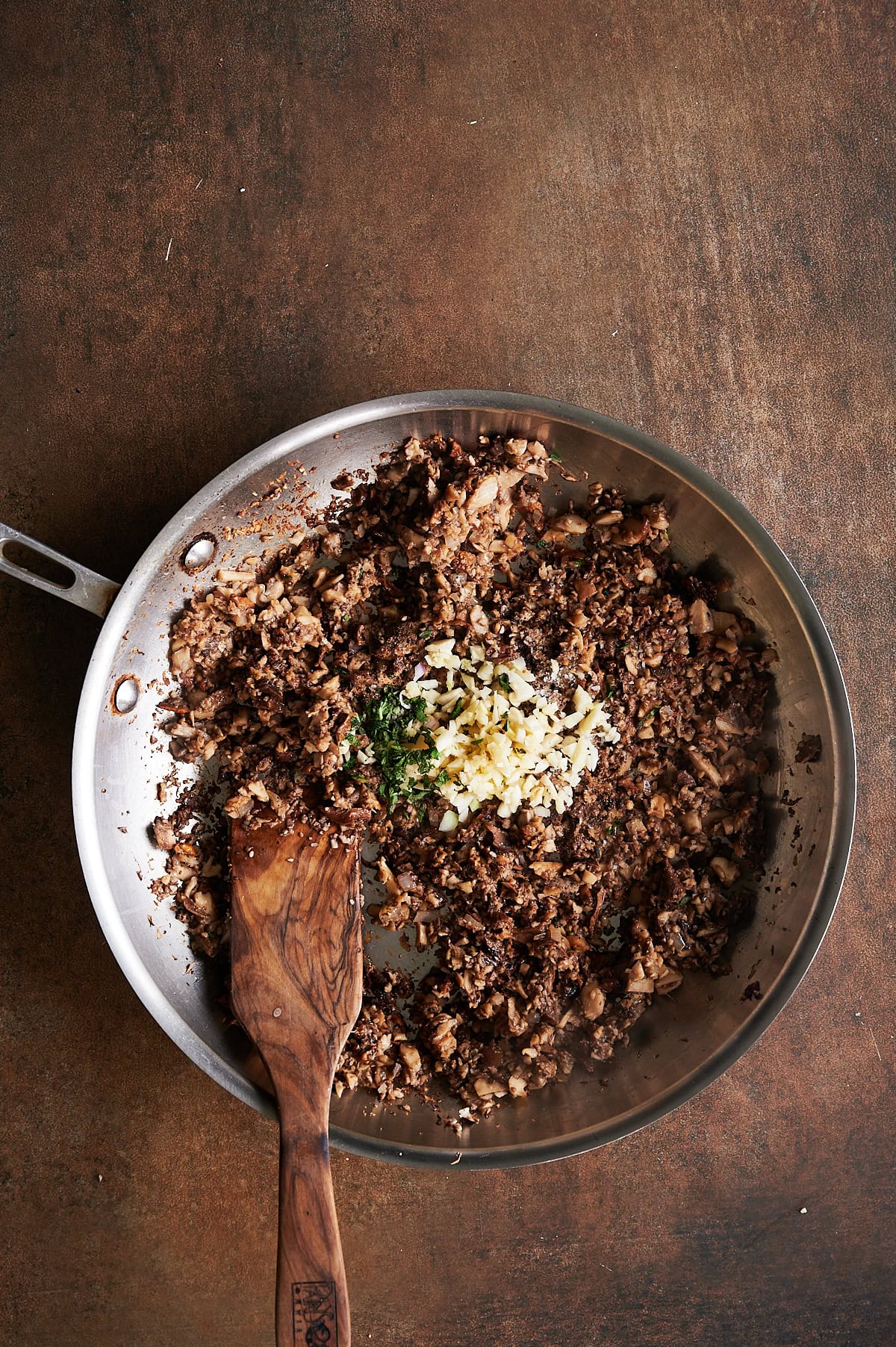 Stainless skillet of chopped mushroom ravioli filling topped with diced garlic and fresh herbs on a brown background.